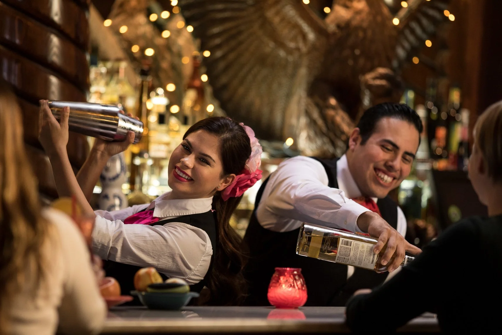 Smiling female bartender in vintage attire pouring whiskey for a male customer at a lively bar with dim lighting and string lights.