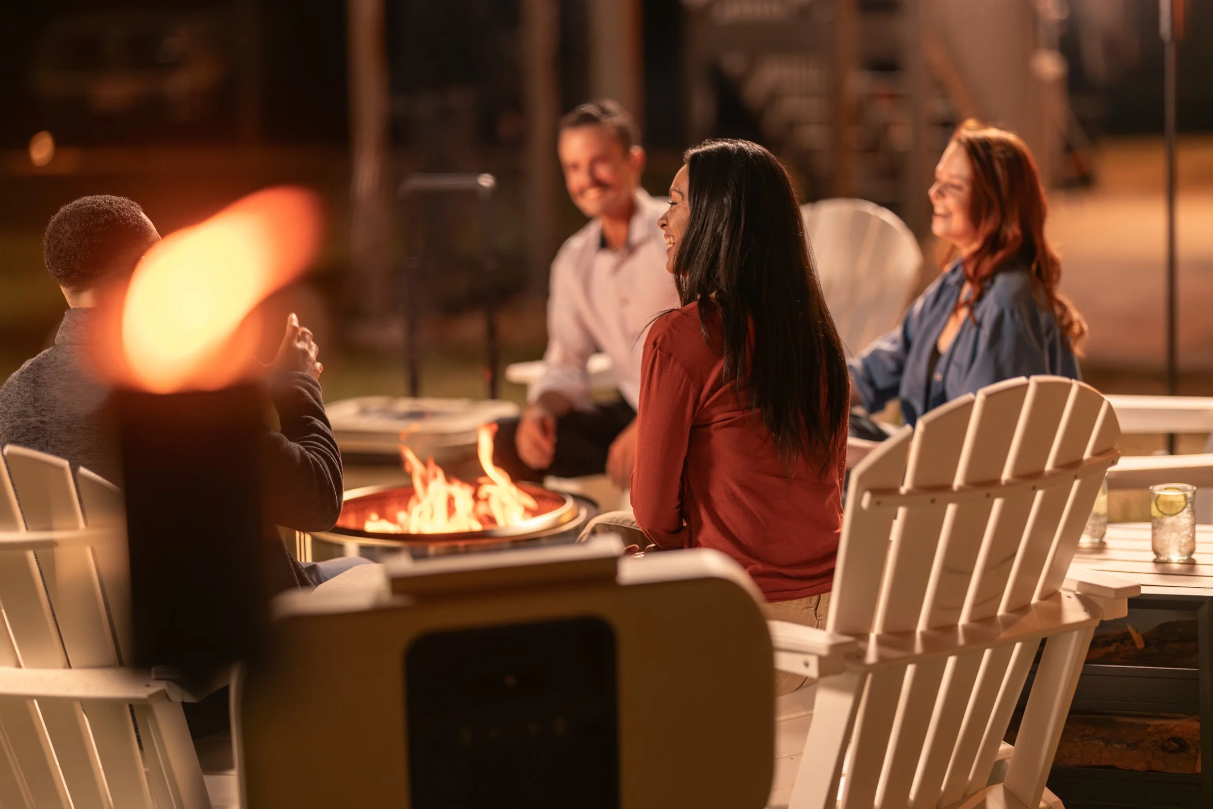 Four people sitting around a fire pit and socializing at night outdoors, smiling and enjoying each other's company.