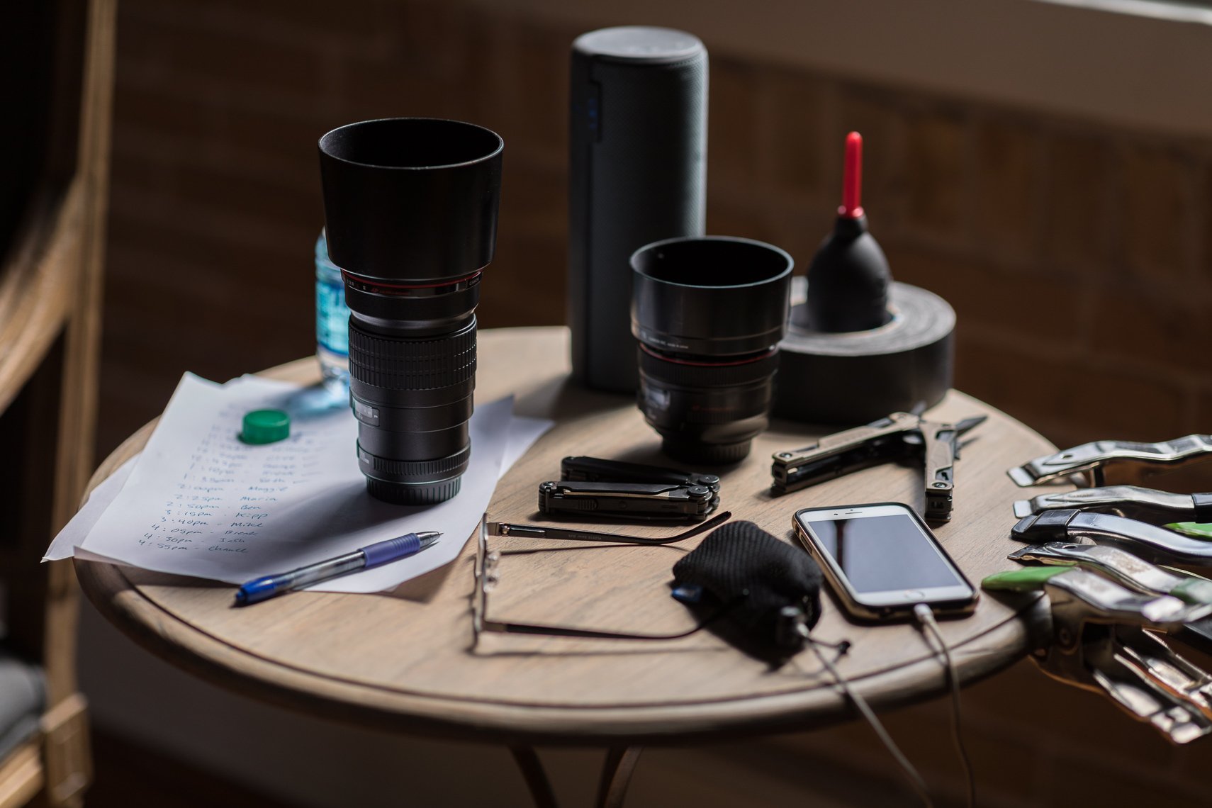 Photograph of a cluttered round wooden table filled with camera lenses, a smartphone, a pen, paper with handwritten notes, tools, a water bottle, a spray bottle, and various small objects in a room with wooden walls.