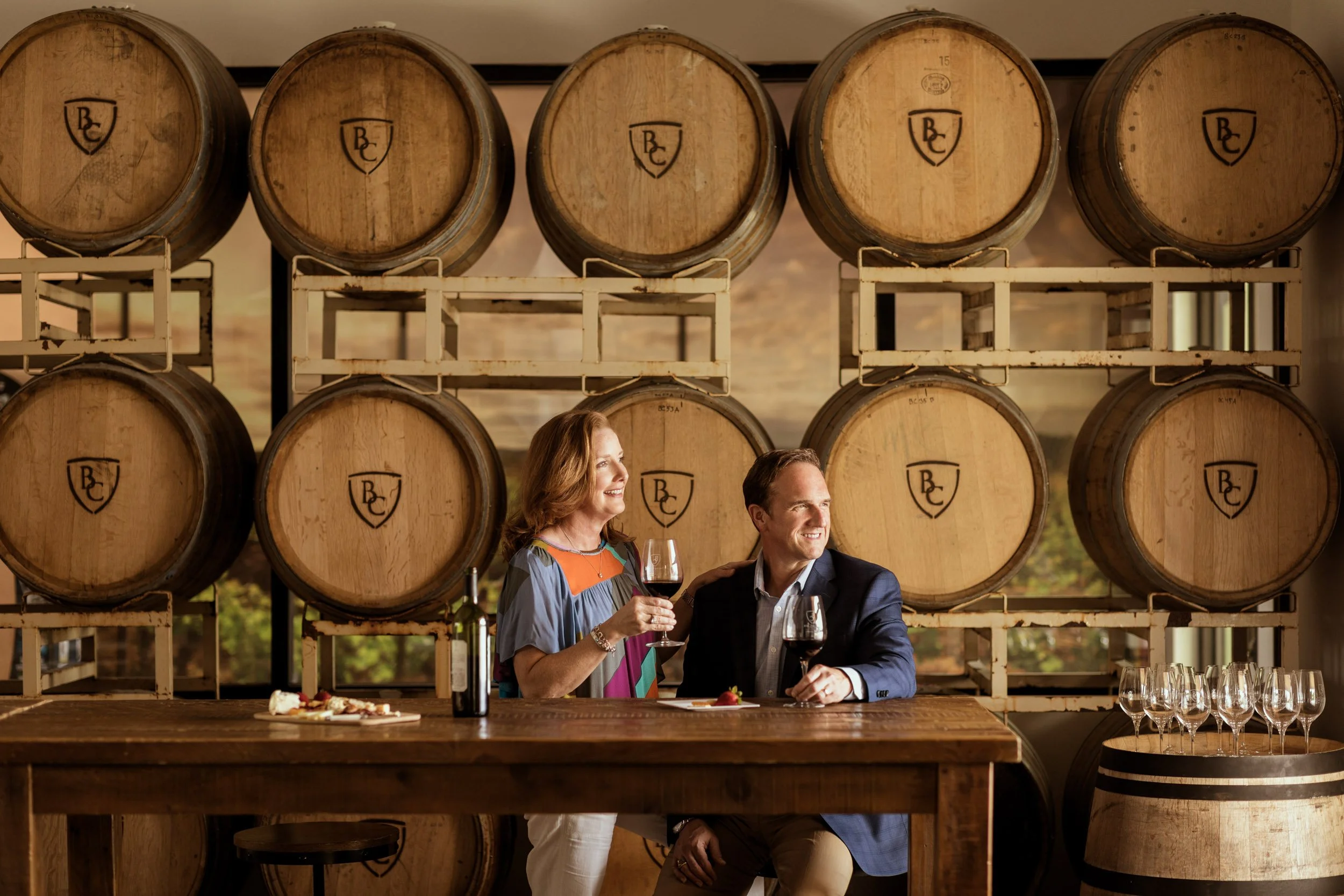 A couple enjoying wine tasting at a winery, standing behind a bar with wine glasses and a plate of food, with large wine barrels in the background.