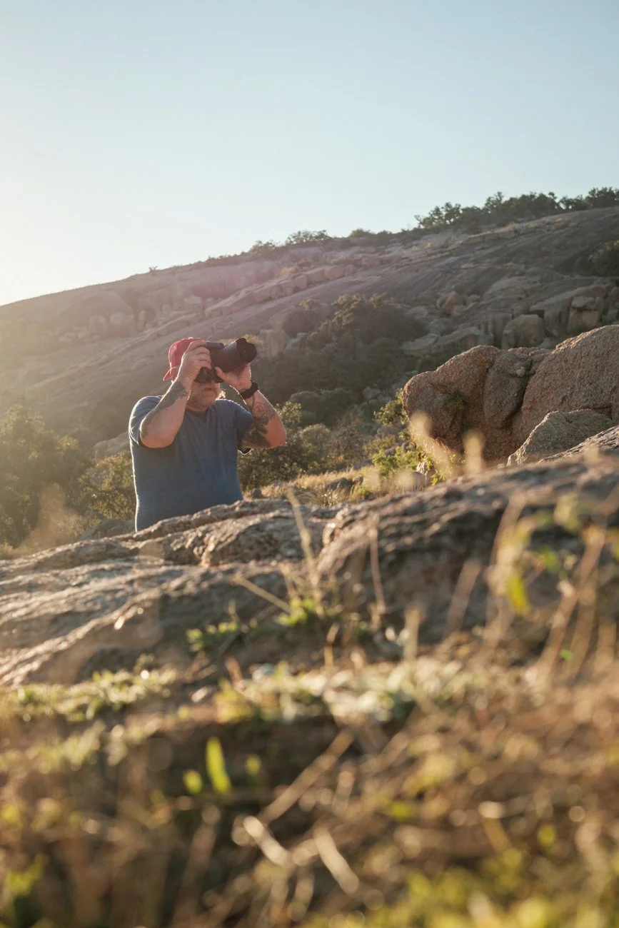 A man in a red cap and gray shirt taking a photograph on rocky terrain during sunset.
