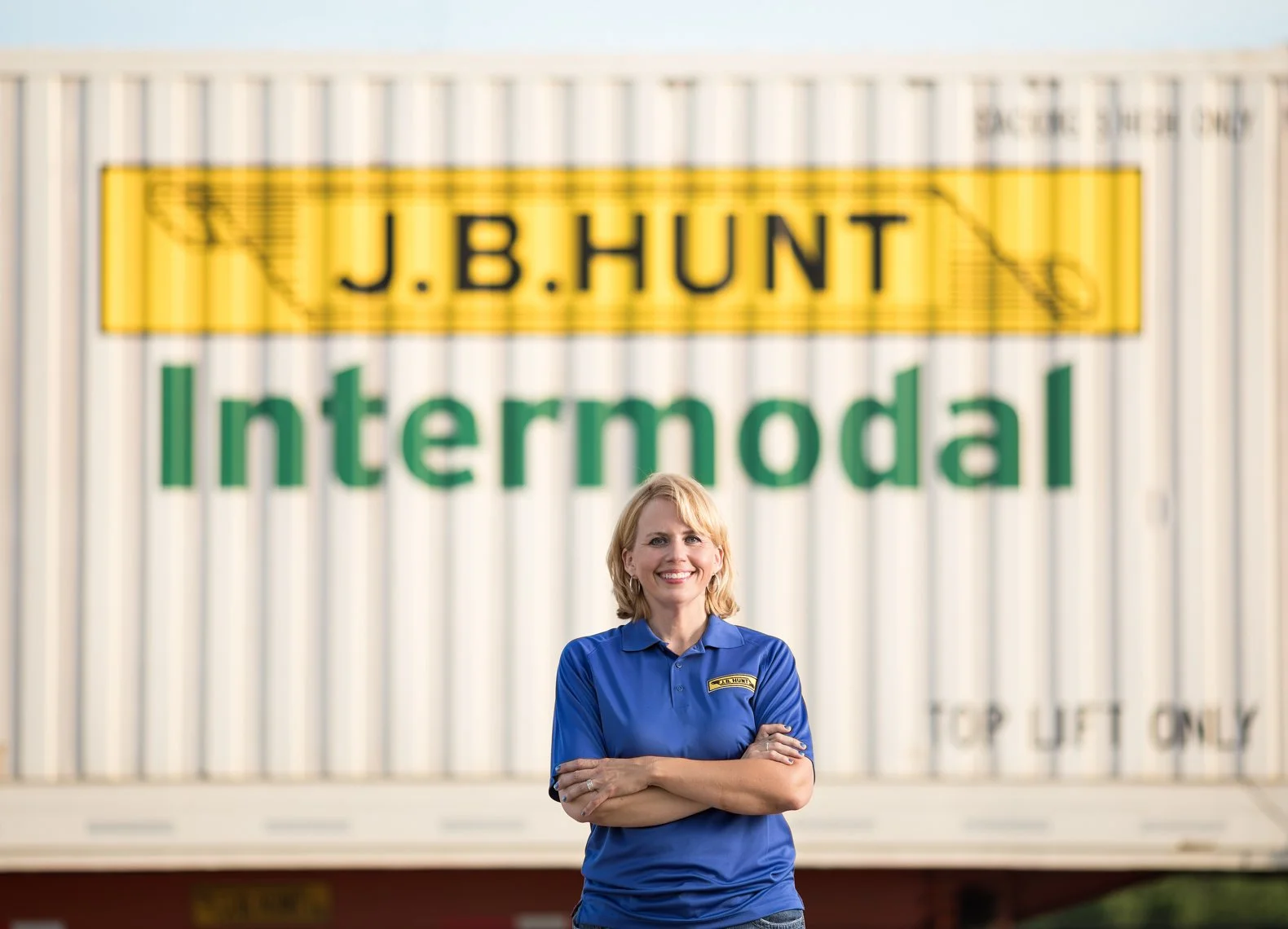 A woman in a blue polo shirt standing with arms crossed in front of a sign that reads 'J.B. Hunt Intermodal' in large green and yellow letters.
