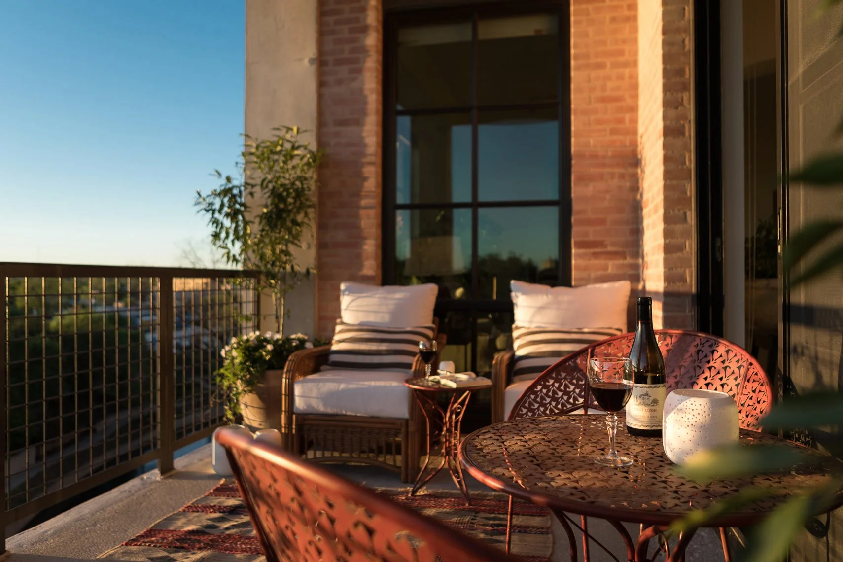 A cozy balcony with two white cushioned chairs with black and white striped pillows, a small round copper table holding a glass of red wine, a bottle of wine, a white decorative candle holder, and a wine opener. The balcony has a brick wall, large bl
