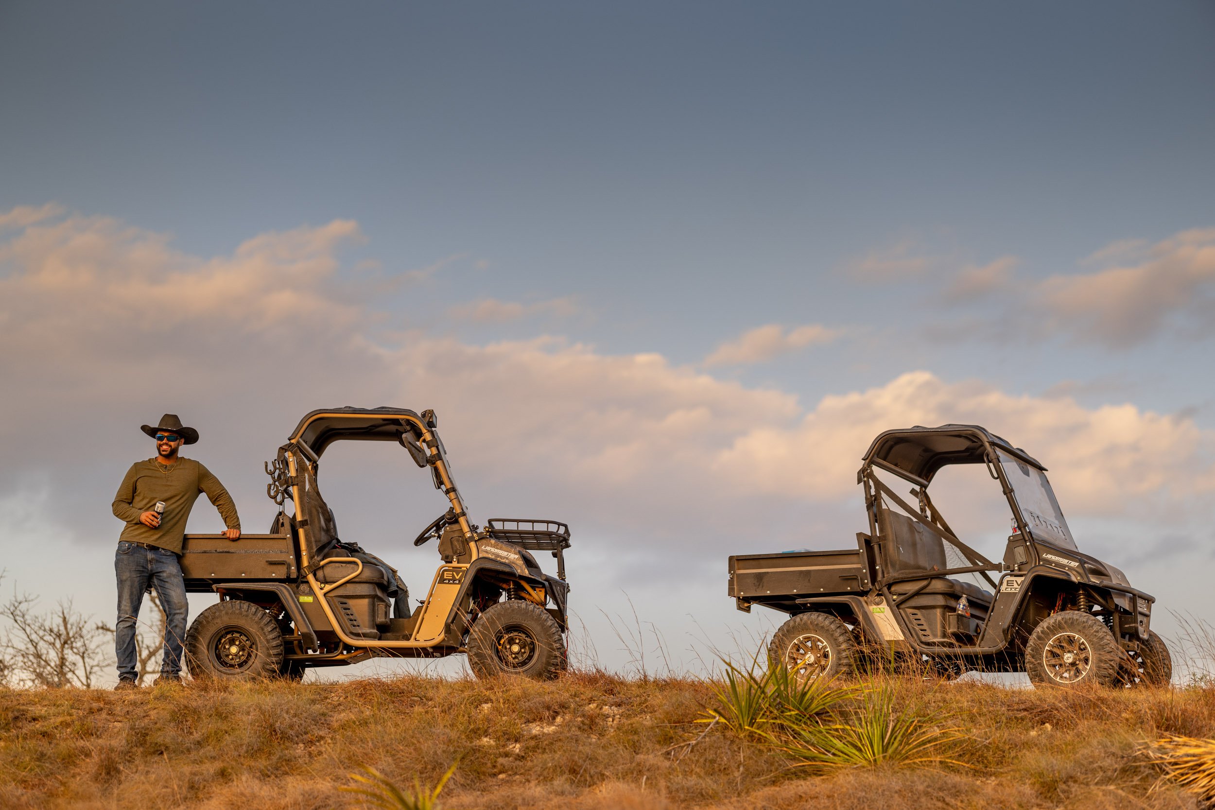 A man wearing a hat and sunglasses standing next to two utility task vehicles (UTVs) on a grassy hill during sunset.