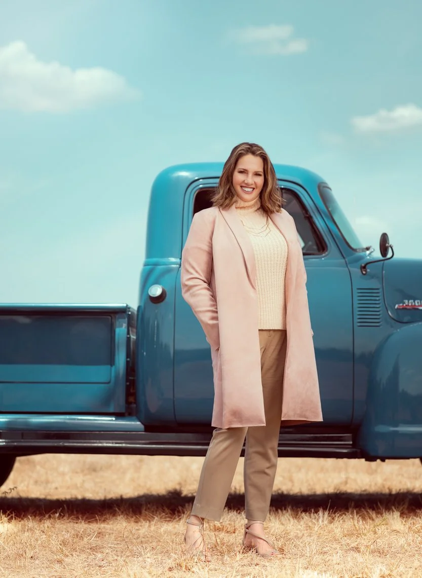 A woman with shoulder-length brown hair wearing a beige coat, light yellow sweater, beige pants, and beige heels, standing in front of a blue vintage truck on a grassy field under a partly cloudy sky.