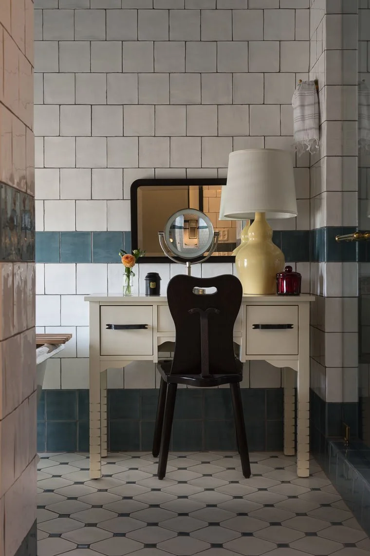 A bathroom vanity table with a large beige lamp, a mirror, a small plant, a candle, and a red jar. A black chair is in front. The wall behind has white and dark green tiles, and there is a white towel hanging on the wall.