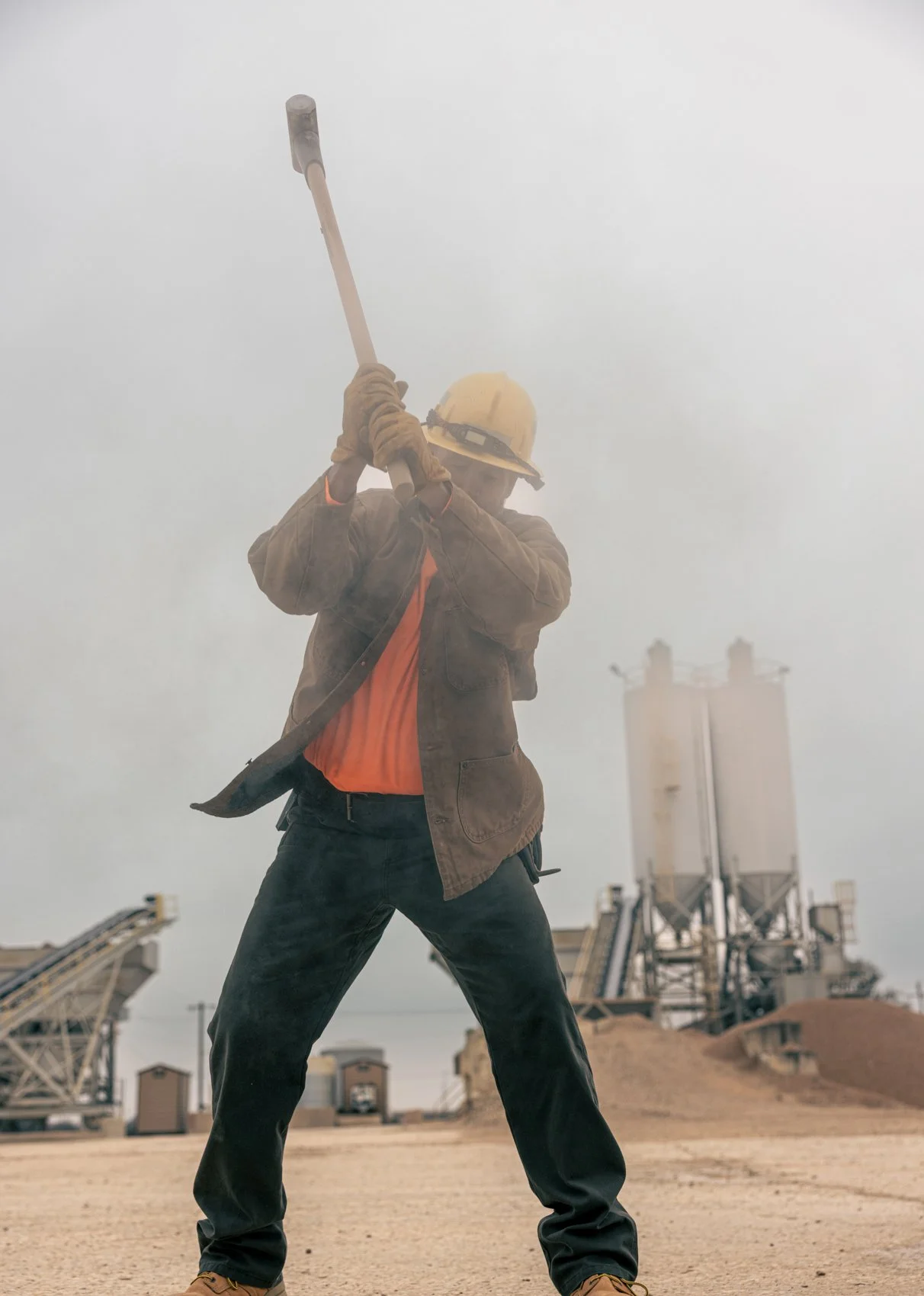 A construction worker wearing a yellow hard hat, brown jacket, orange shirt, black pants, and gloves, swinging a sledgehammer at a construction site with factory buildings and conveyor belts in the background.