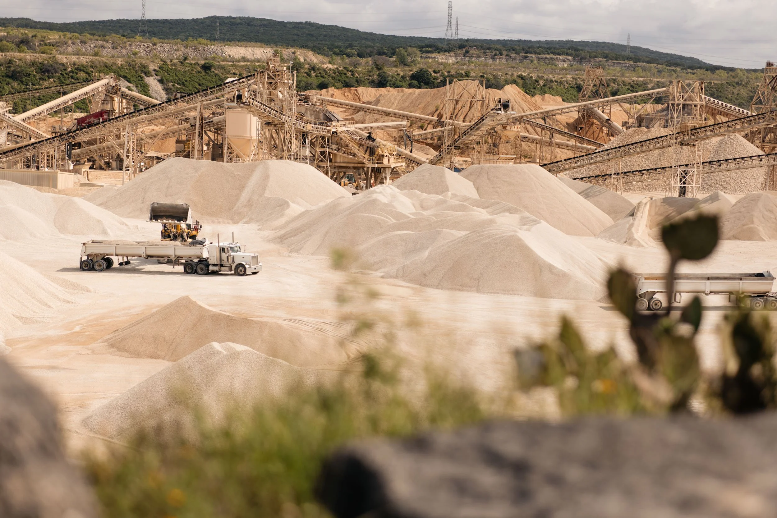 Open-pit sand quarry with large piles of sand, conveyor belts, and mining equipment in a rural area with hills and power lines in the background.