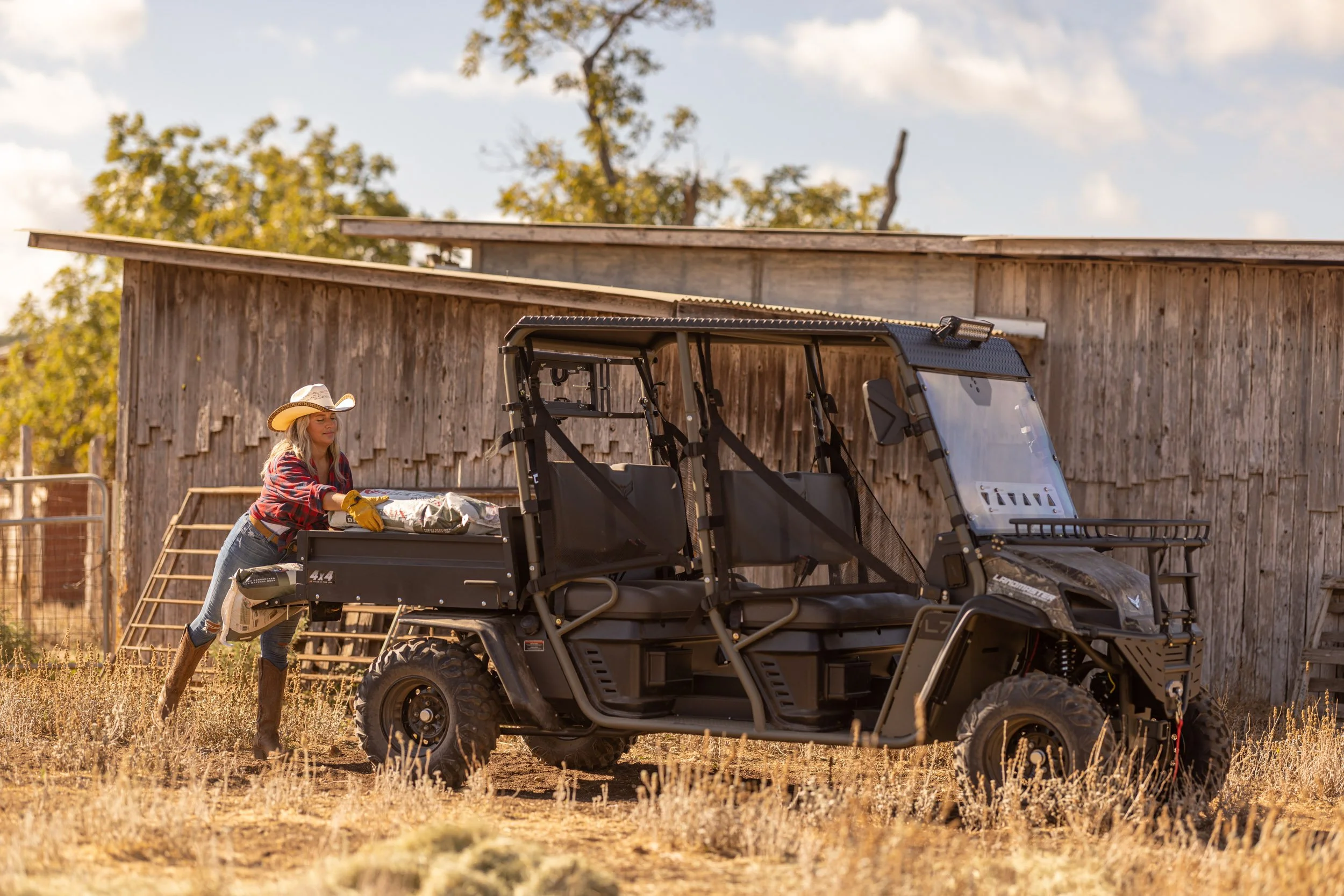 A woman with long blonde hair wearing a cowboy hat, red plaid shirt, and jeans loading supplies into a black utility vehicle in a rural farm setting with a wooden barn and trees in the background.