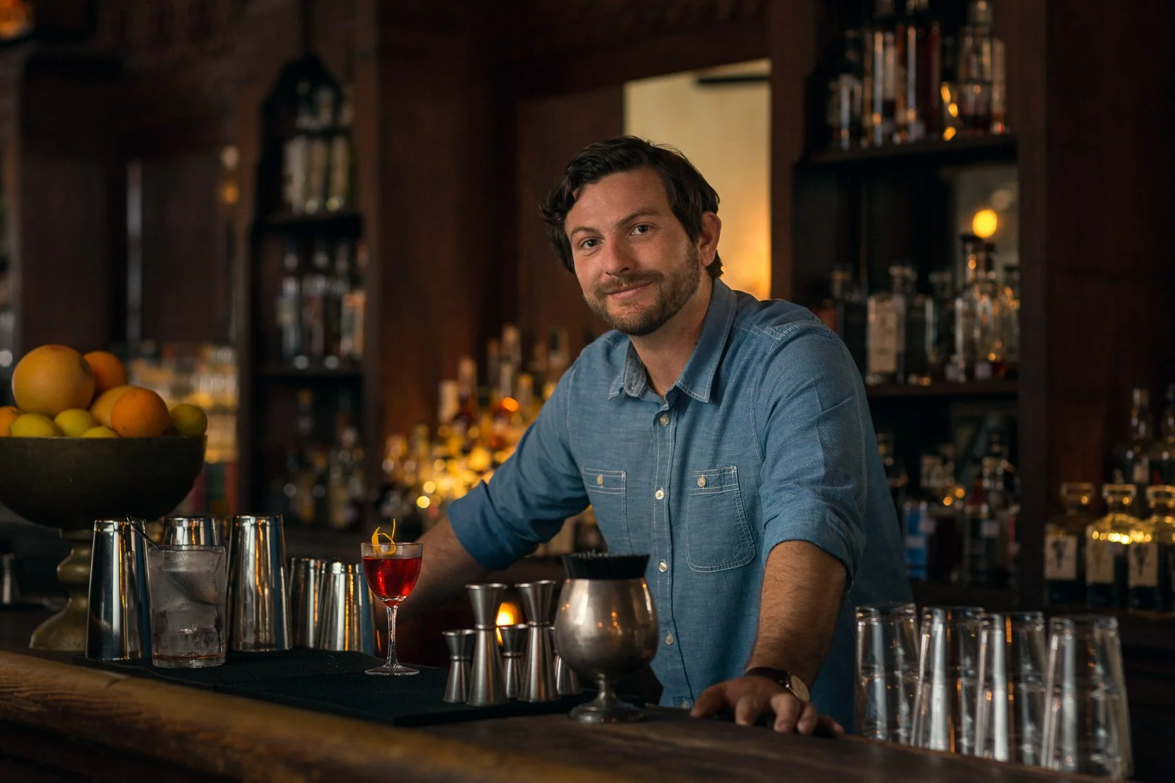 A man with dark hair and a beard, wearing a blue denim shirt, sitting behind a bar counter with various bar tools, a bowl of fruit, and glasses, smiling at the camera in a dimly lit bar with shelves of liquor bottles in the background.
