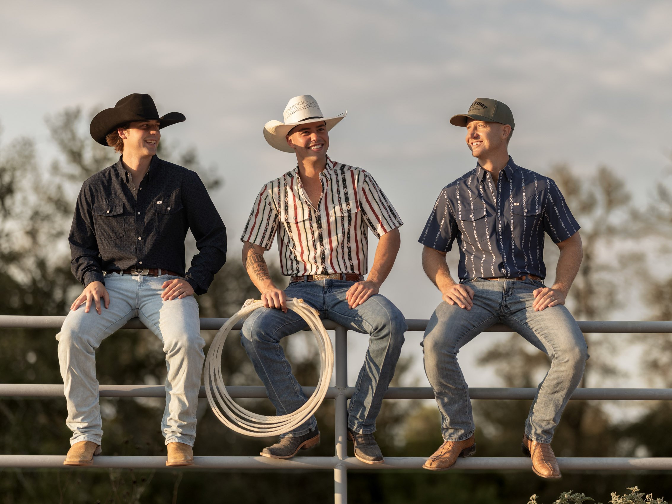 Three men sitting on a fence, dressed in western attire with cowboy hats, smiling and talking outdoors against a background of trees and a cloudy sky.