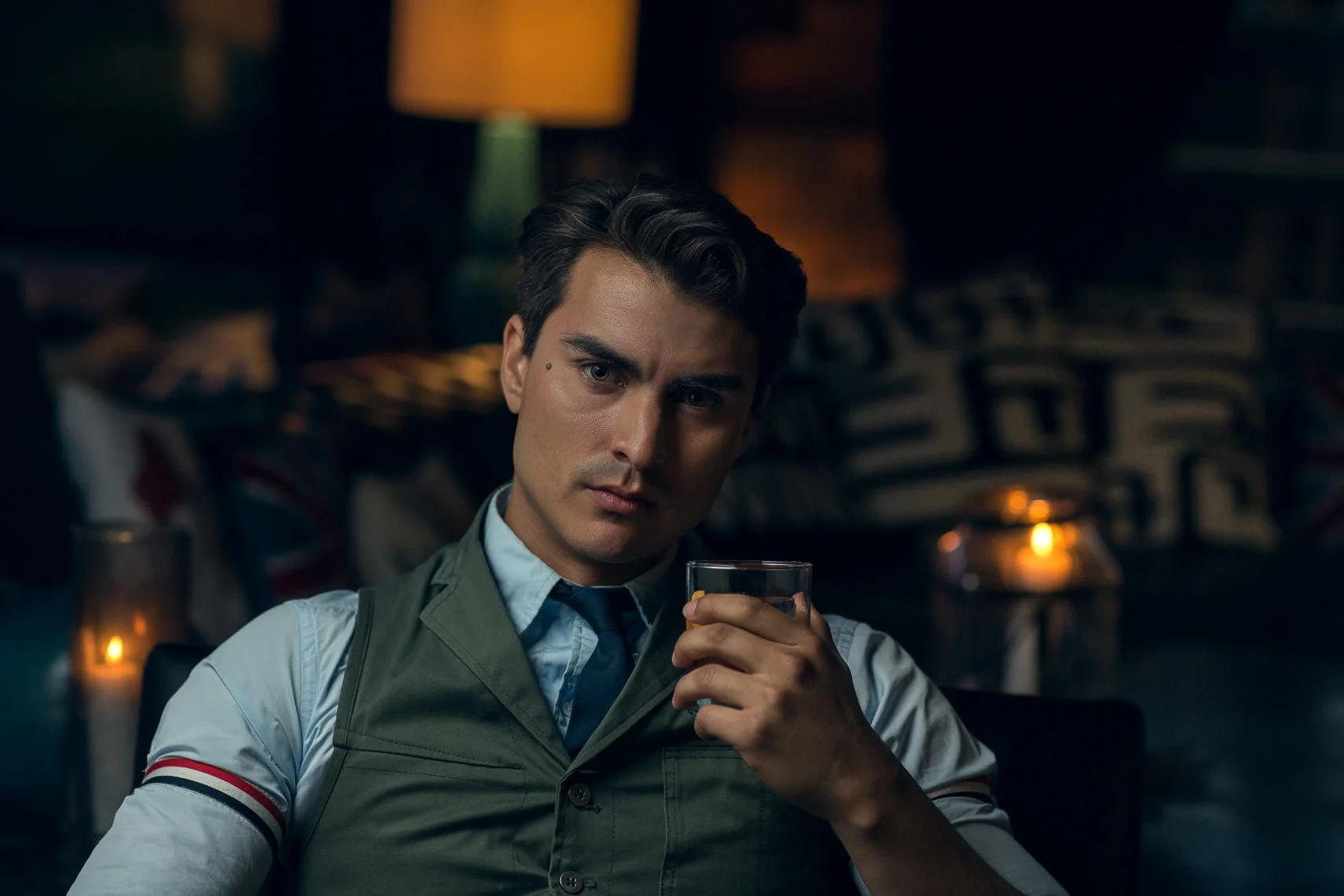 A young man with dark hair, wearing a light blue shirt, tie, and a military-style vest, is sitting in a dimly lit room, holding a glass of drink, looking directly at the camera.