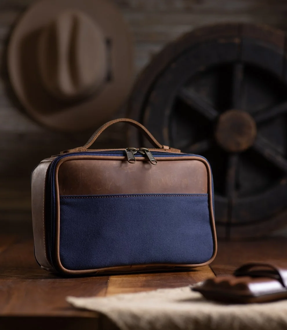 A brown and navy blue travel bag on a wooden surface with a vintage wagon wheel and a hat hanging on the wall in the background.