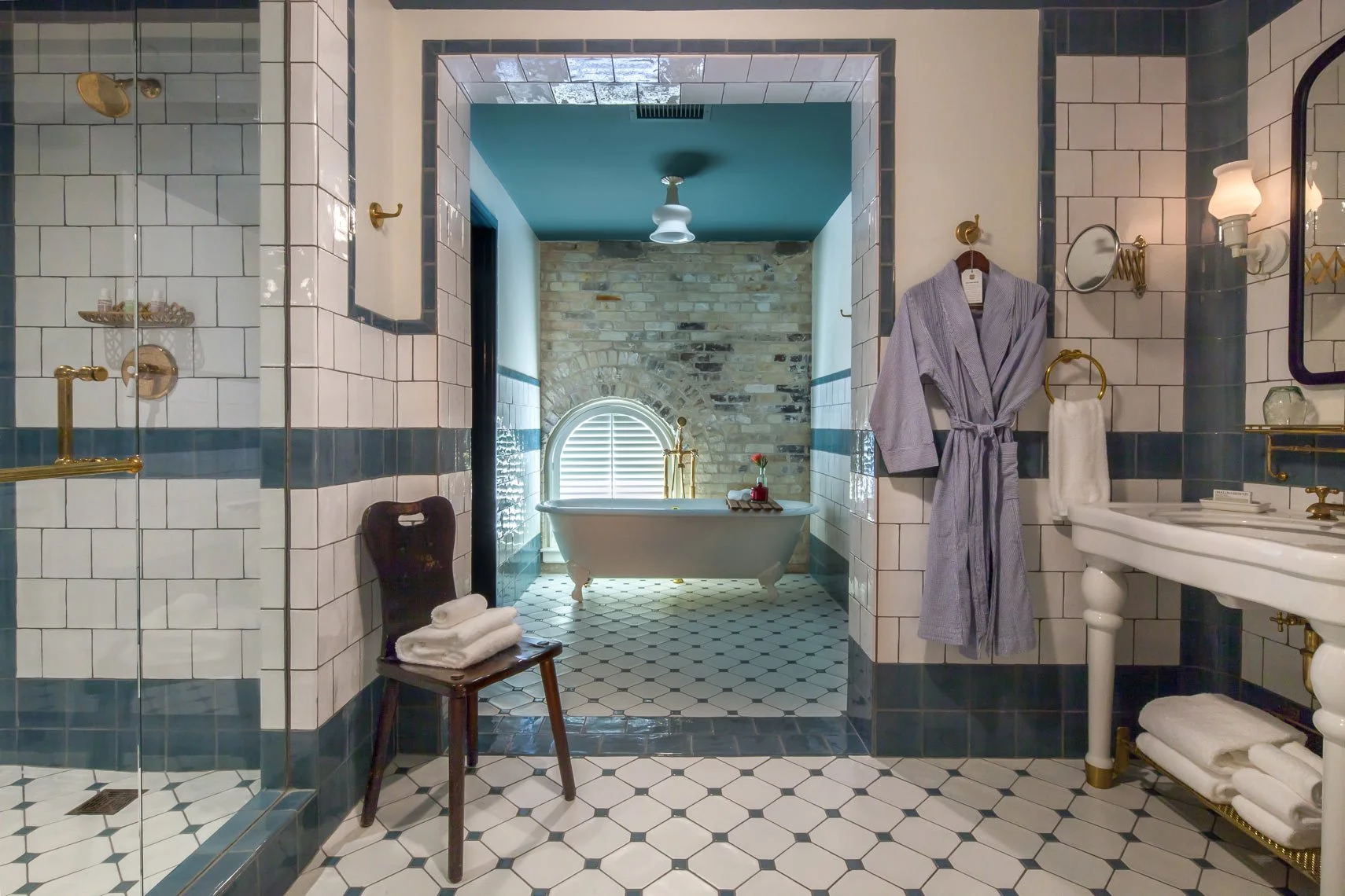 Bathroom with a clawfoot bathtub under a window with shutters, a tiled shower area, a white sink with gold fixtures, a bathrobe hanging on the wall, and a chair with folded towels