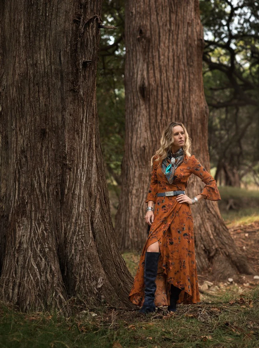 A woman in a rust-colored floral dress with a high slit, wearing black boots, stands in a forest next to tall trees, with her hand on her hip and looking to the side.