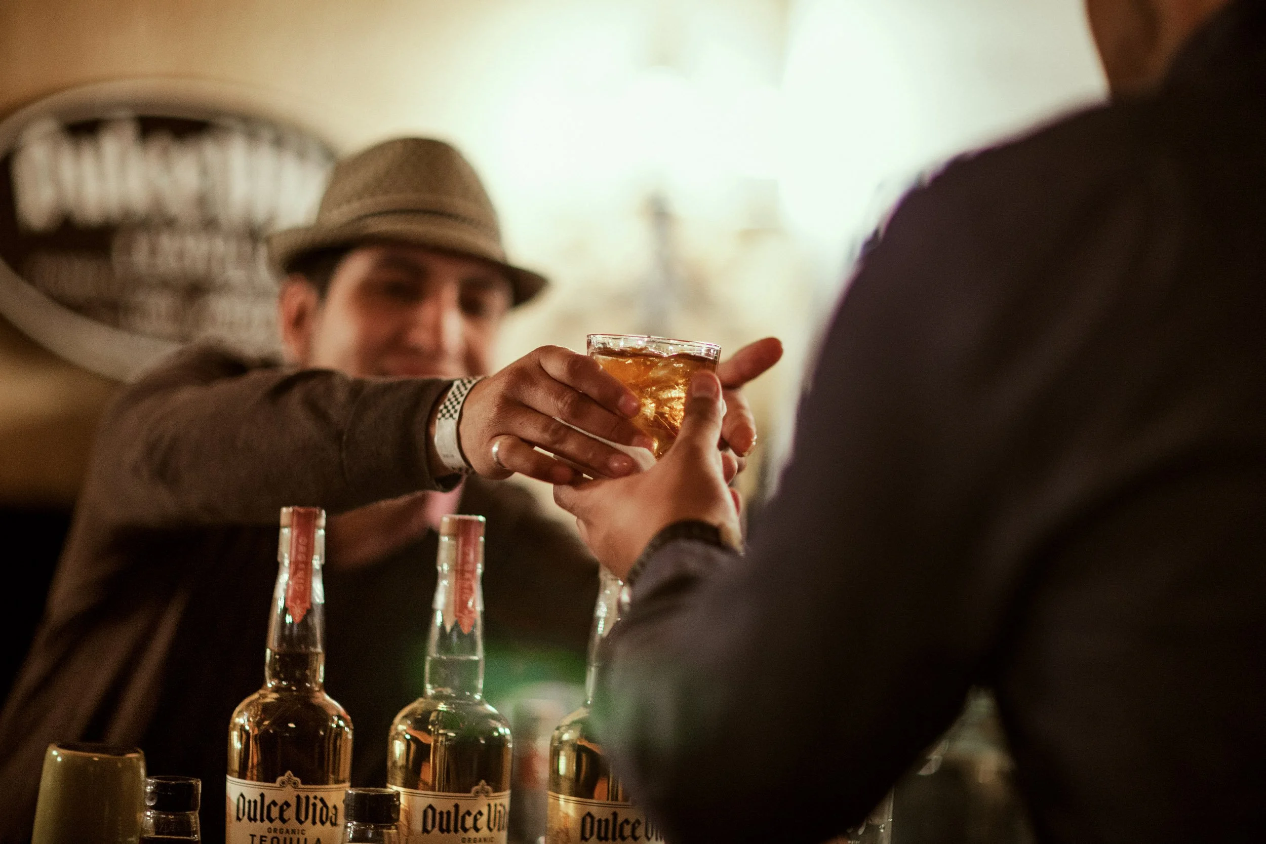 Person in a hat smiling and holding a glass of amber-colored drink, with bottles of Dulce Vida organic tequila in the foreground.