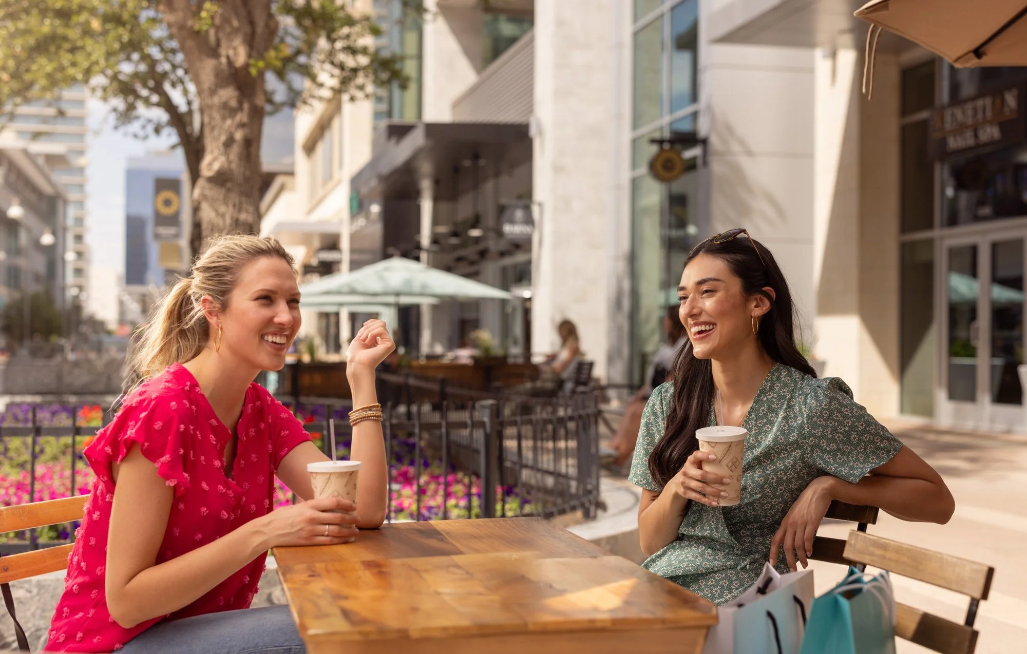 Two women sitting at an outdoor table at a cafe, smiling and having a conversation. One woman is wearing a red patterned dress and the other is in a green floral dress, each holding a paper cup with a drink. The background shows a city street with tr
