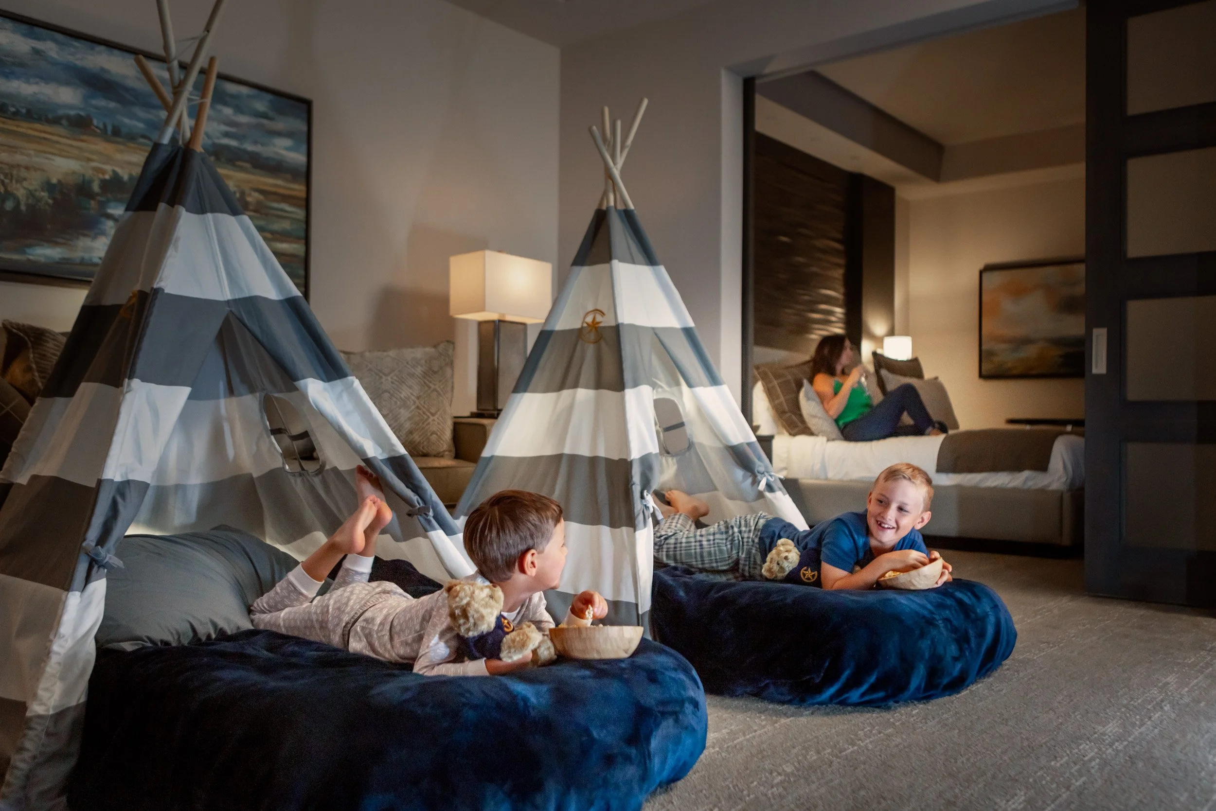 Children playing and relaxing on the floor inside a hotel room, with two small tents set up, a woman sitting on the bed in the background, and artwork on the walls.