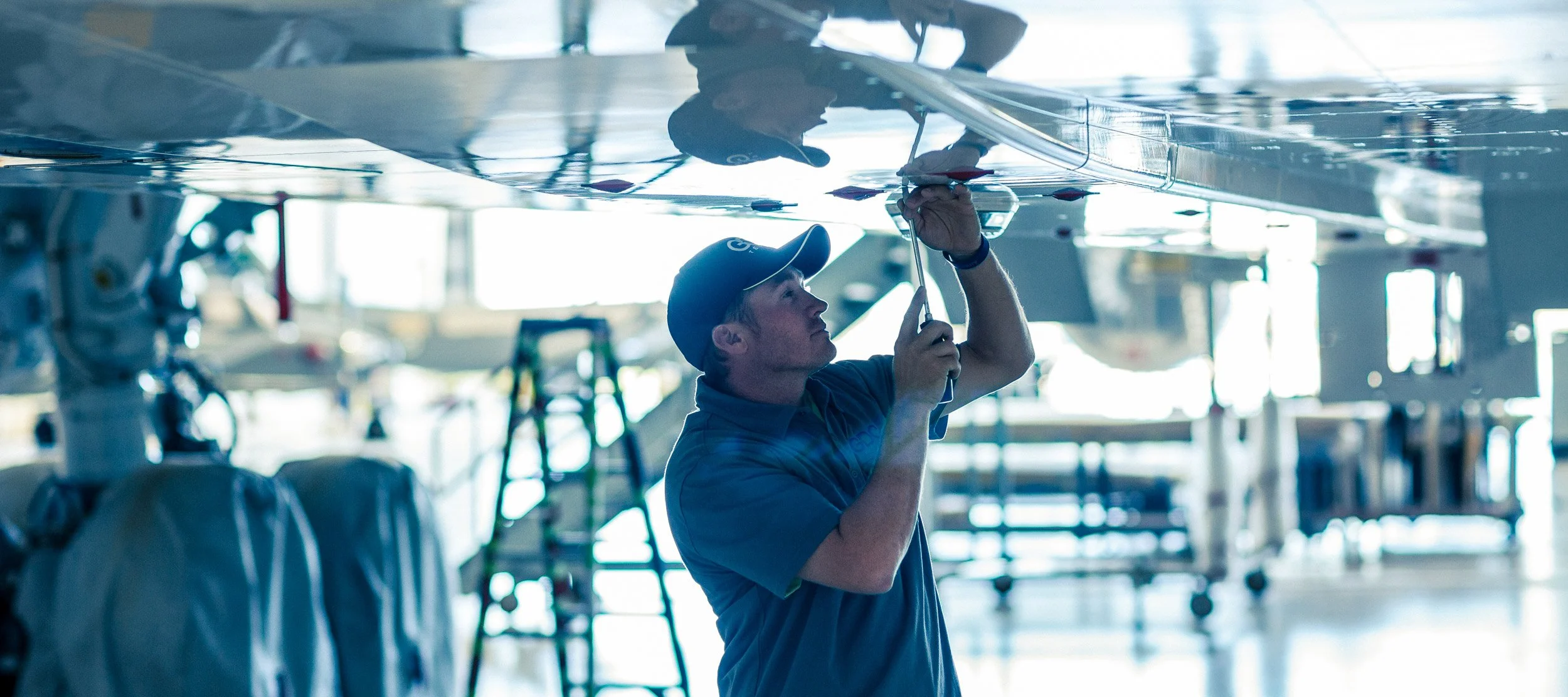 A man in a warehouse or hangar, working on the underside of an aircraft, using a tool.
