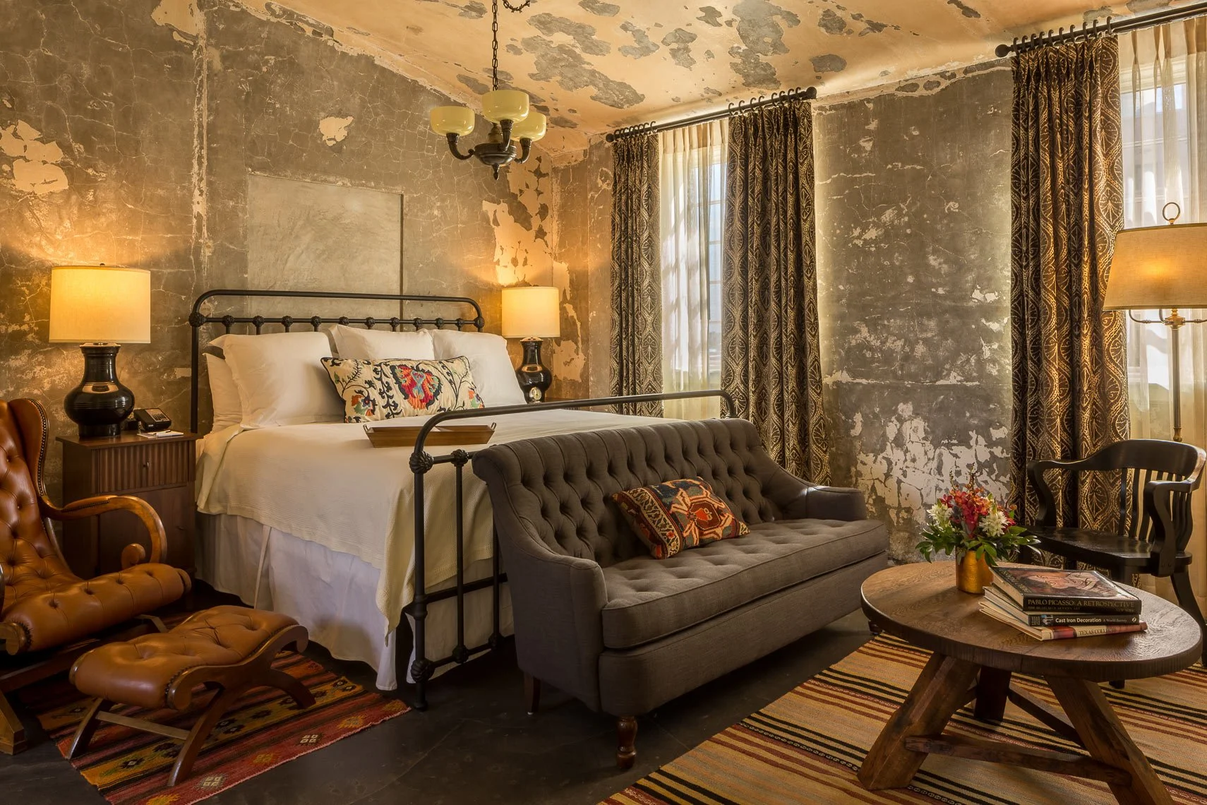 A cozy bedroom with a distressed concrete wall, a vintage bed with white bedding and a decorative pillow, a side table with a lamp, a brown tufted leather chair, a gray tufted sofa with a decorative pillow, a round wooden coffee table with a flower a