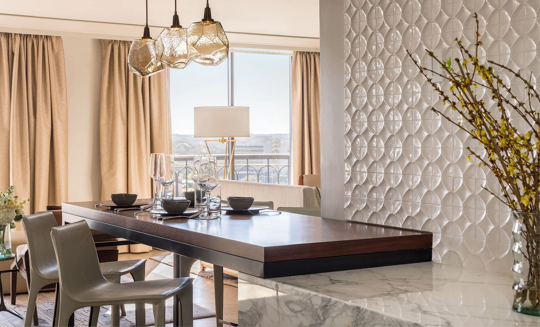 Interior of a modern dining room with a dark wood dining table set with black bowls and glasses, beige chairs, beige curtains, a marble countertop, a white textured wall, a floor lamp, and a window with a city view.