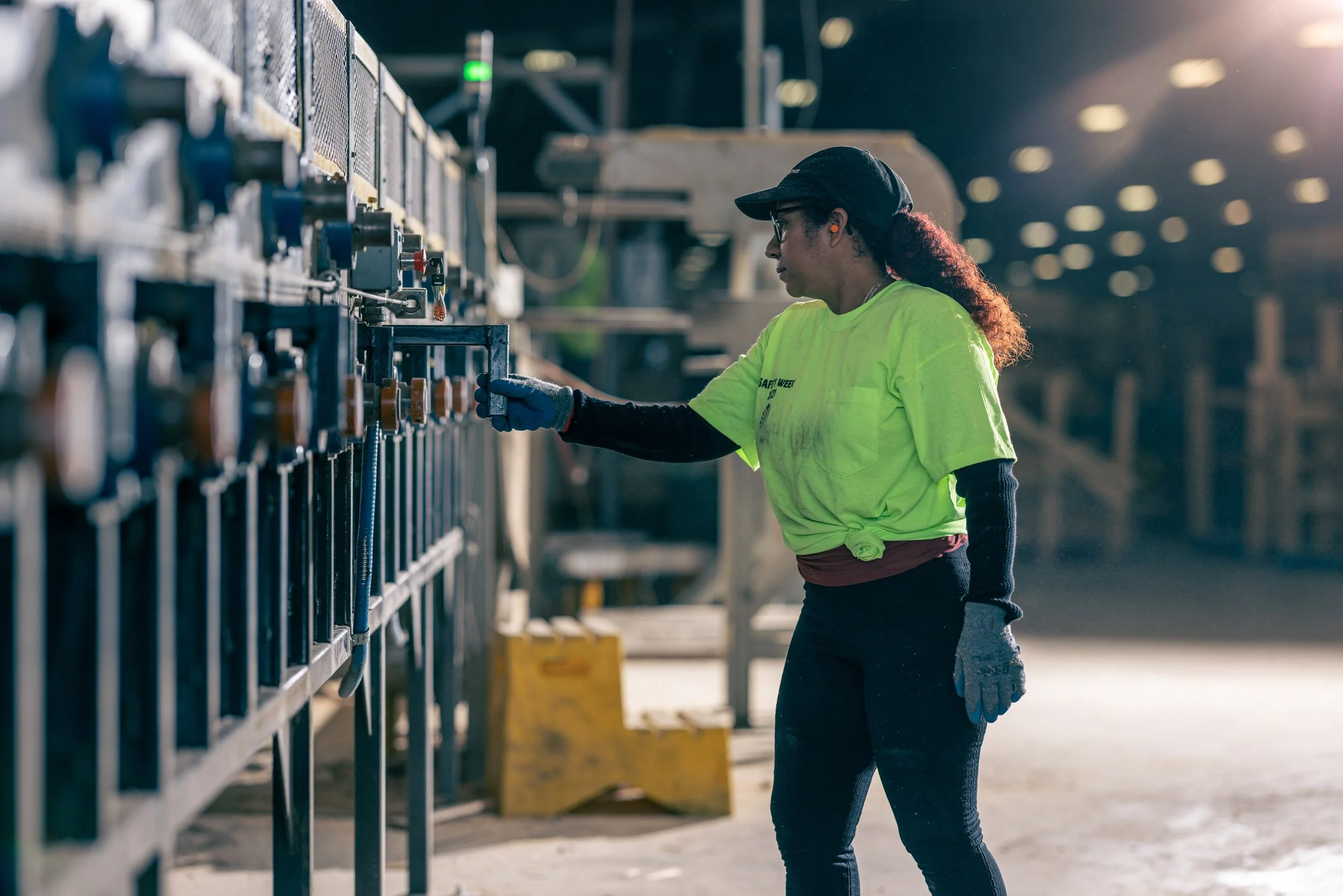 A woman wearing a neon yellow safety shirt and black cap working with machinery outdoors, illuminated by bright sunlight.