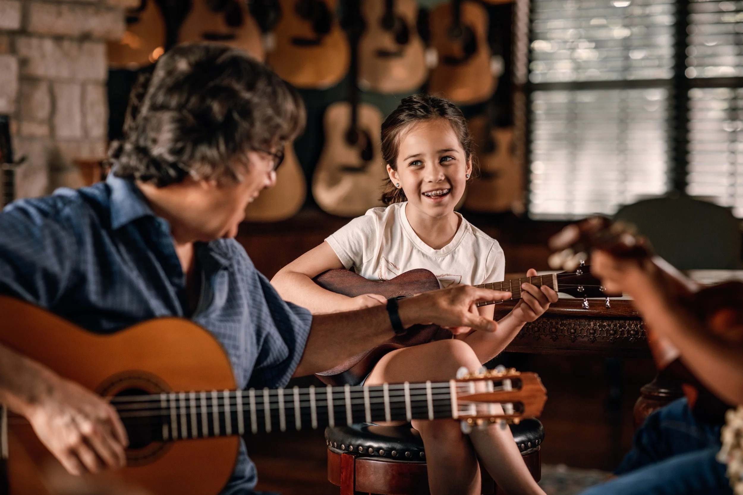 A girl happily playing the guitar while her older friend, also with a guitar, shares a moment of music and joy together in a cozy room with guitars hanging on the wall in the background.