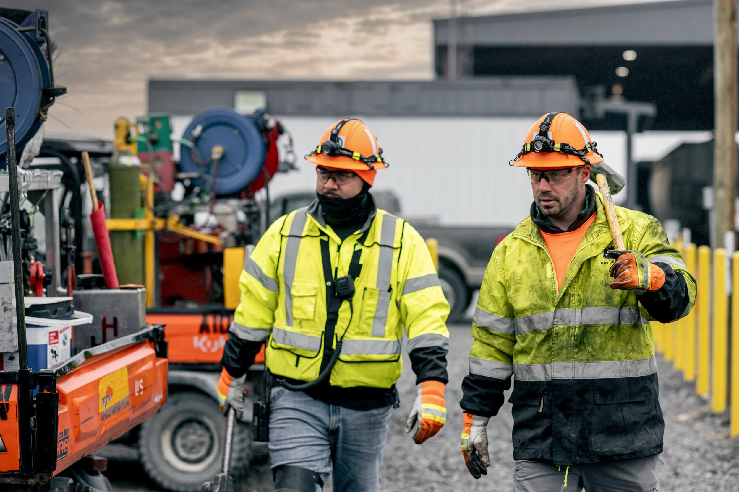 Two construction workers in yellow safety jackets, orange helmets, and safety glasses walking outdoors on a construction site with equipment and buildings in the background.