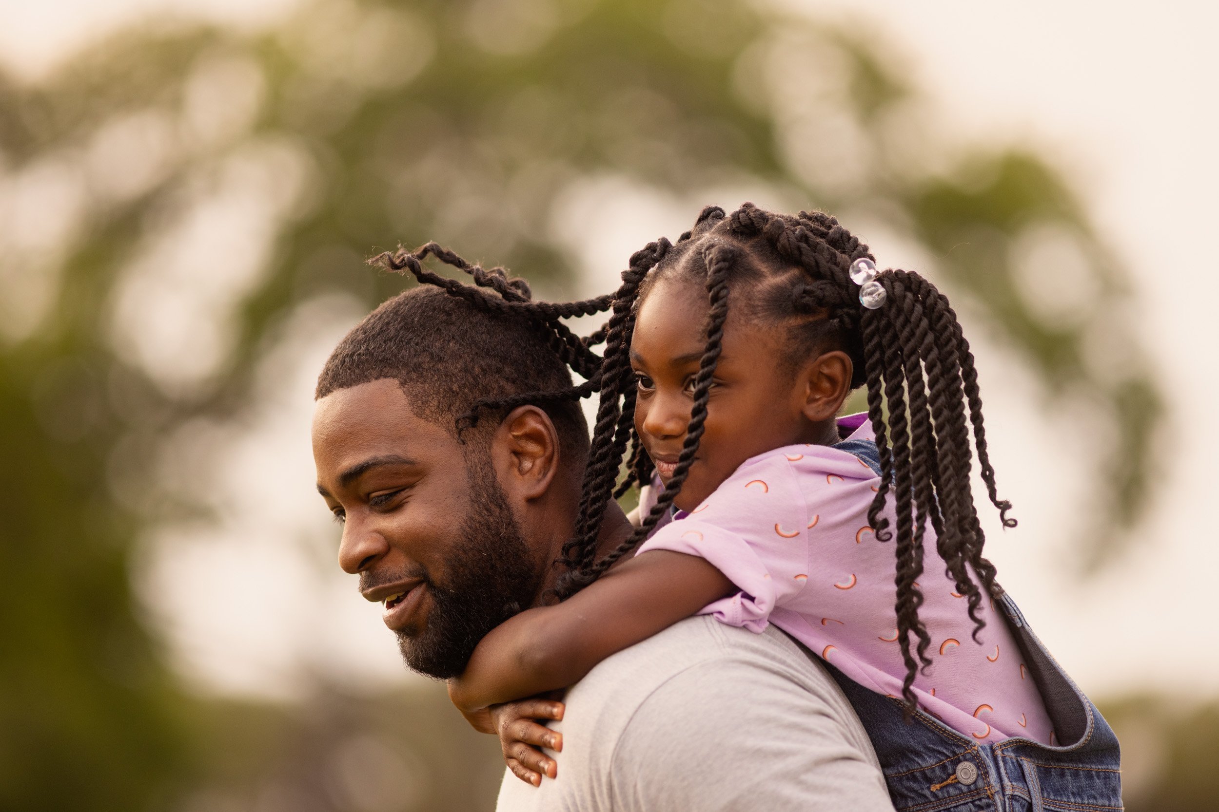 A man carrying a young girl on his shoulders outdoors during daytime with trees in the background.