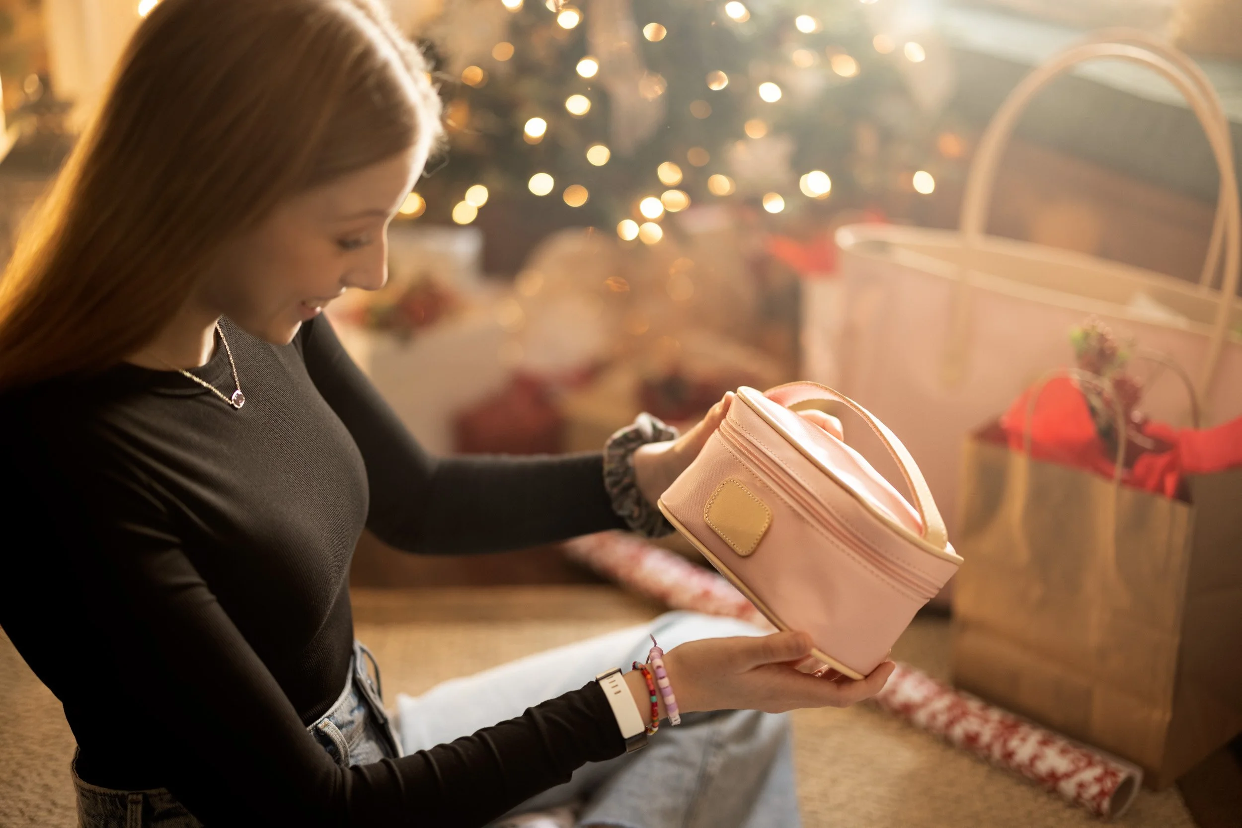 A young woman with long brown hair looking into a pink gift bag during Christmas, with a decorated Christmas tree and lit ornaments in the background.