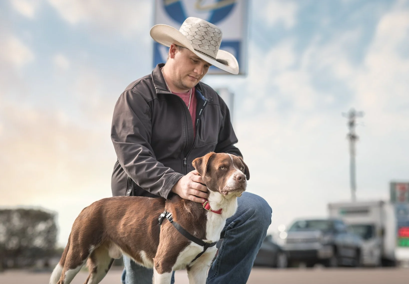 A man wearing a cowboy hat and a black jacket is kneeling down and gently holding a brown and white dog on a leash, in a parking lot with cars and cloudy sky in the background.
