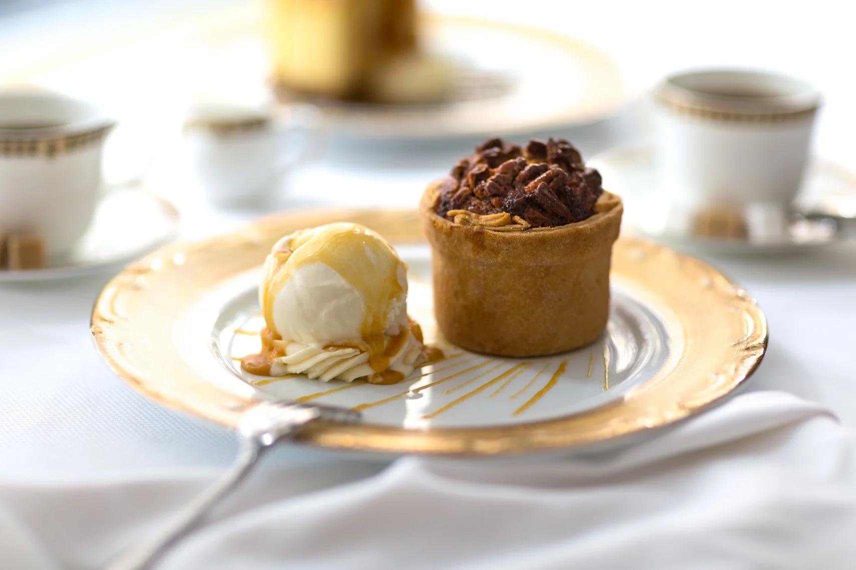 A dessert plate with a scoop of vanilla ice cream drizzled with caramel sauce and a chocolate chip muffin, set on a fancy gold-rimmed plate with a teaspoon in front, and a blurred background of tea cups and saucers.