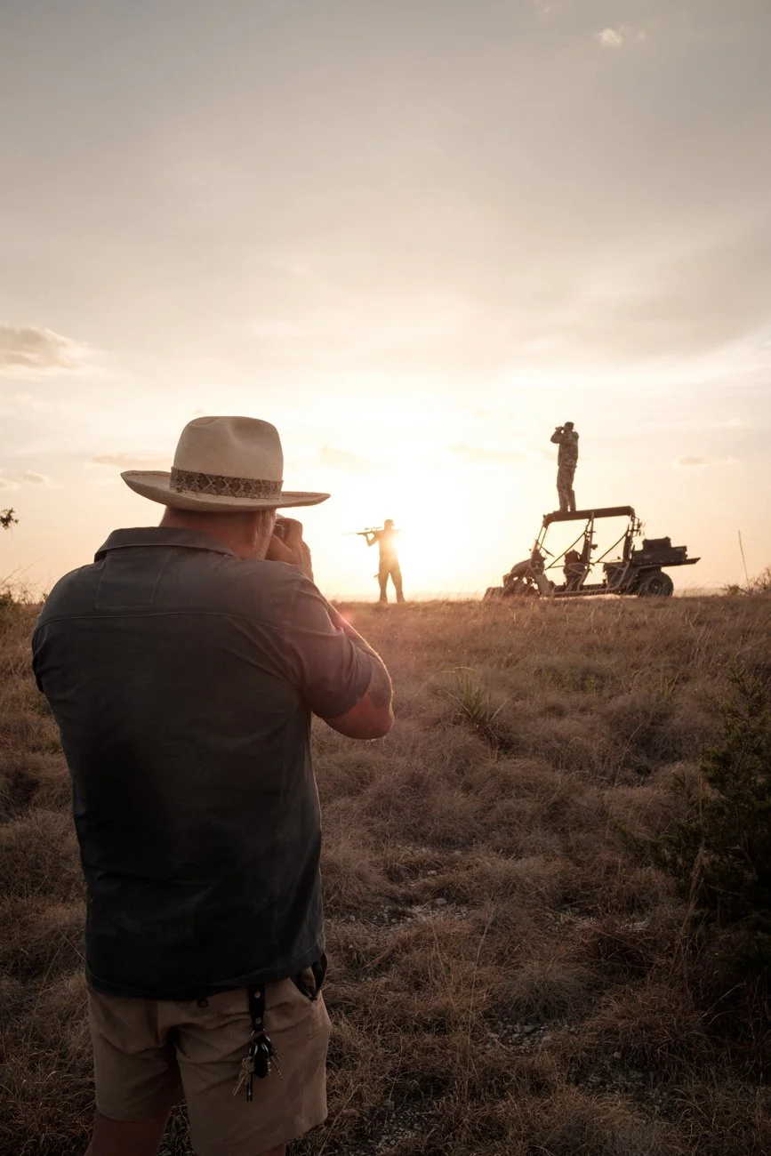 A person wearing a hat and casual clothes is taking a photo or video of two soldiers in a rural field at sunset. One soldier is aiming a rifle, while the other is standing on a small off-road vehicle, looking through binoculars or a camera.