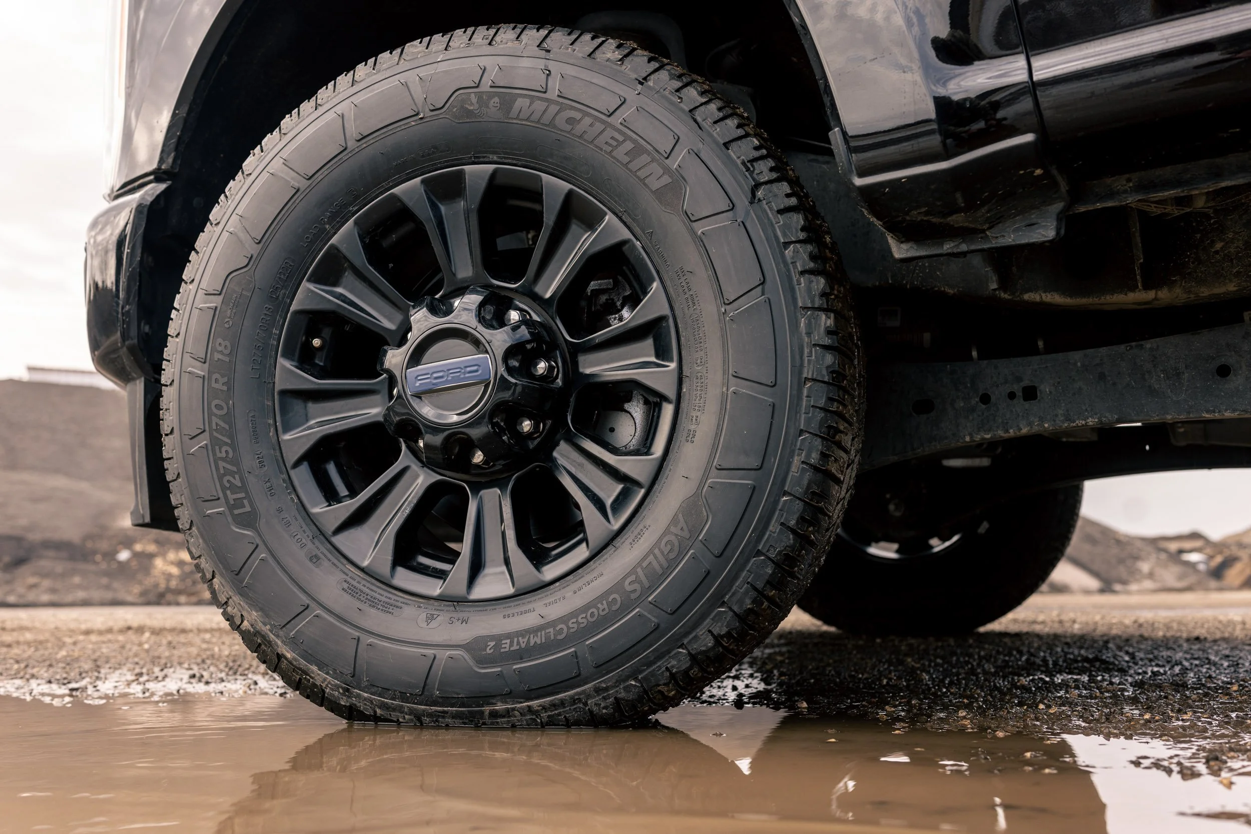 A close-up of a black Ford vehicle tire on a gravel surface with water pooling underneath, and part of the tire and wheel well visible.