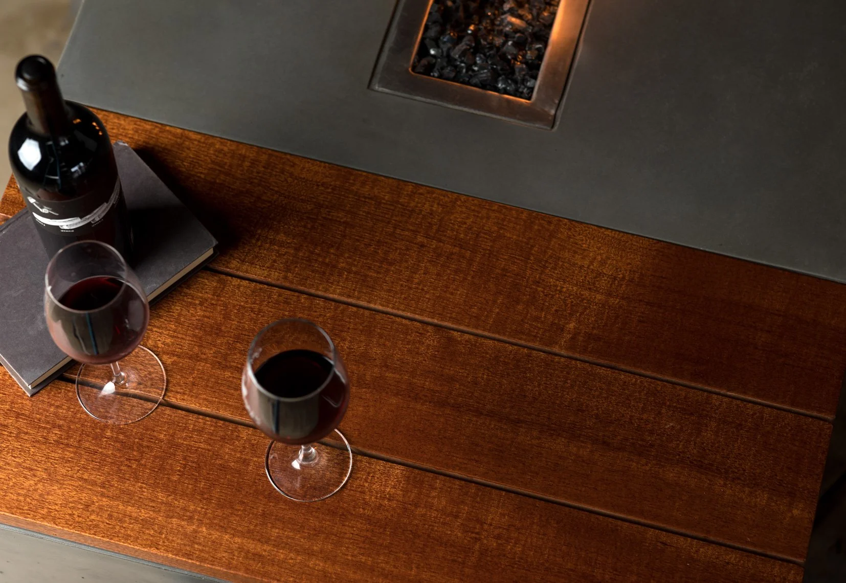 Three glasses of red wine on a wooden table with a bottle of wine, two closed notebooks, a black book, and a fireplace with black stones.