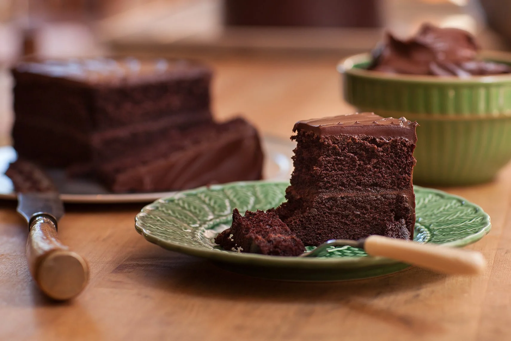 A slice of chocolate cake on a green decorative plate with a fork, with more chocolate cake in the background and a knife on a wooden surface.