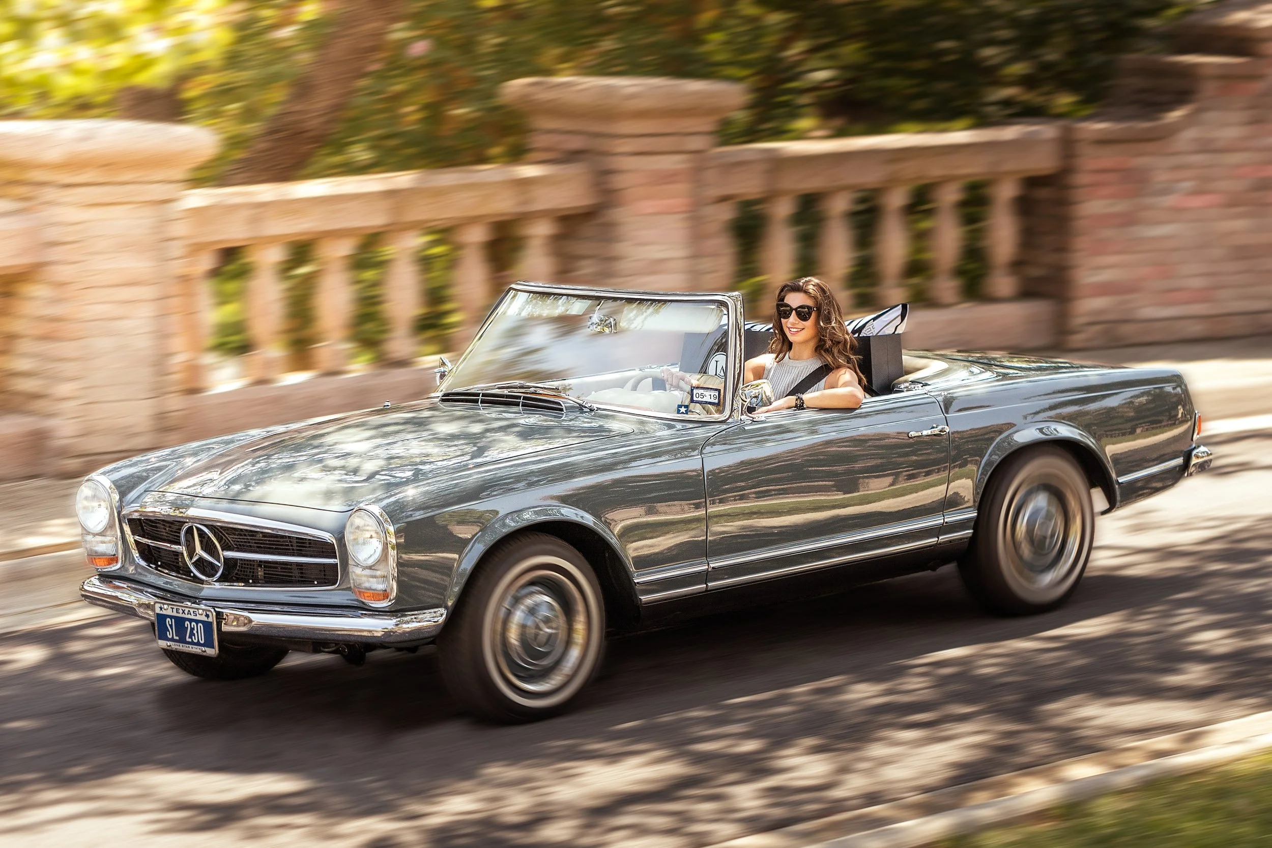 A woman in sunglasses driving a classic silver Mercedes-Benz convertible with a Texas license plate along a street with a brick and stone fence and trees in the background.