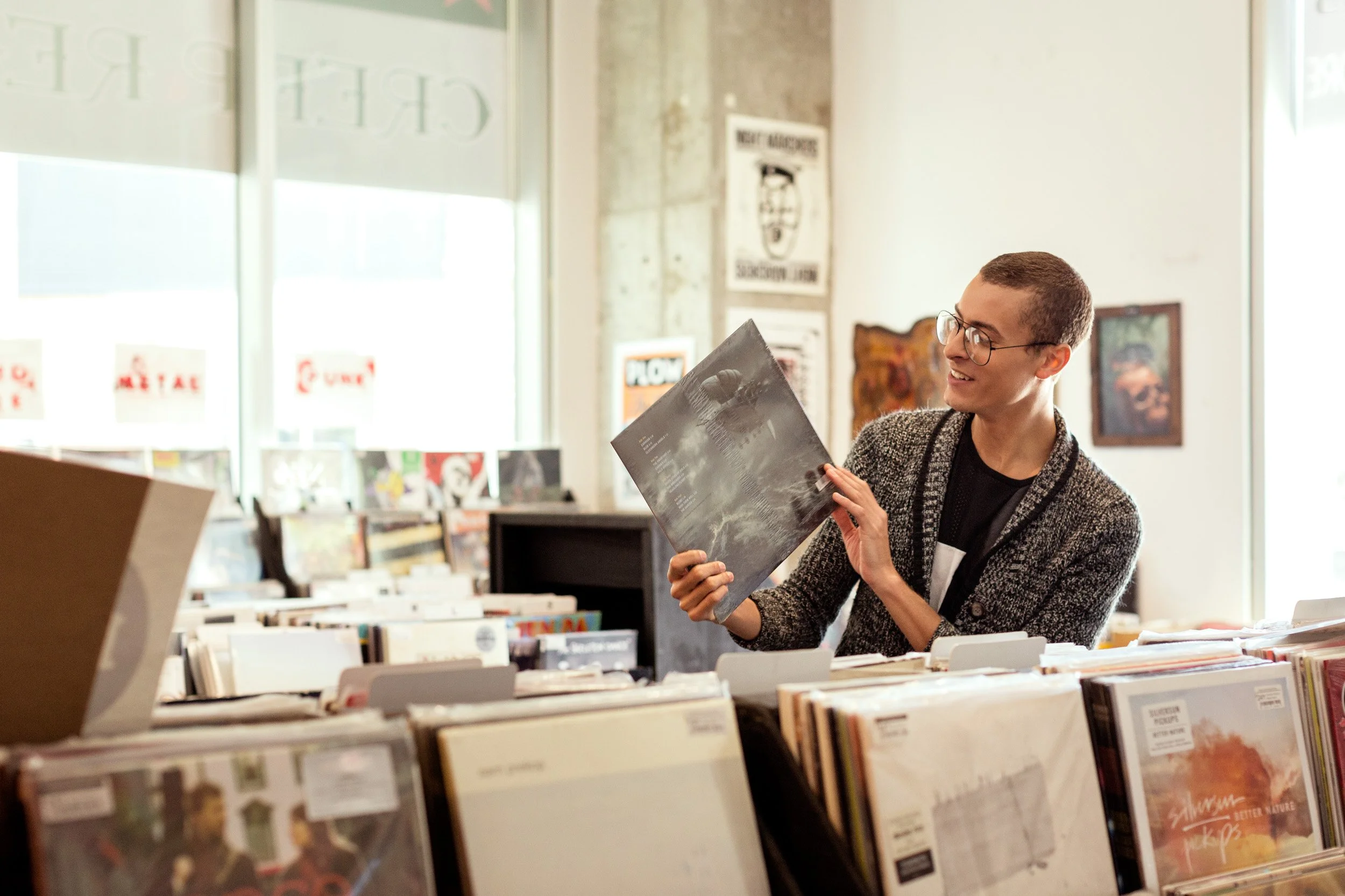 A person with glasses and a black shirt smiling while looking at a vinyl record in a record store with shelves of records and posters on the wall.