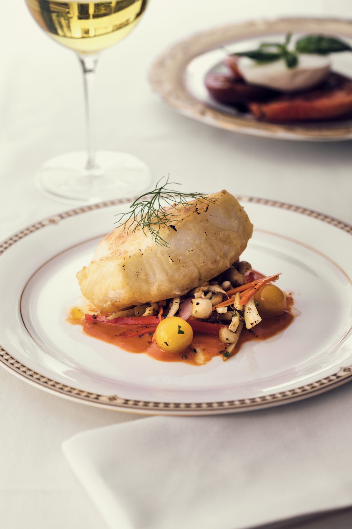 A plated piece of fish with herbs and vegetables, served with sauce, garnished with a sprig of dill on a decorative plate, with a glass of white wine and a side dish in the background.