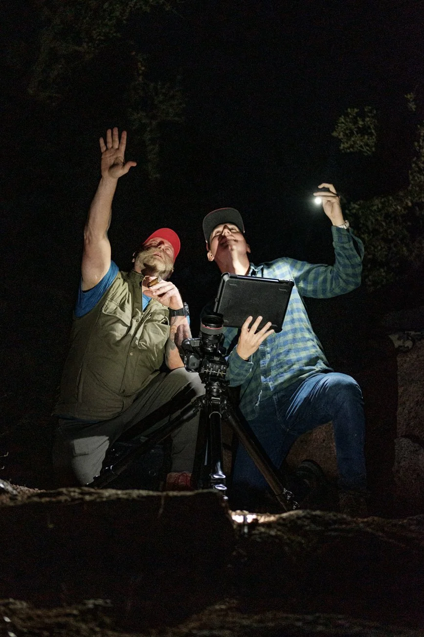 Two men in outdoor clothing at night, using a camera and tablet to observe the sky, with one holding a flashlight, surrounded by trees.