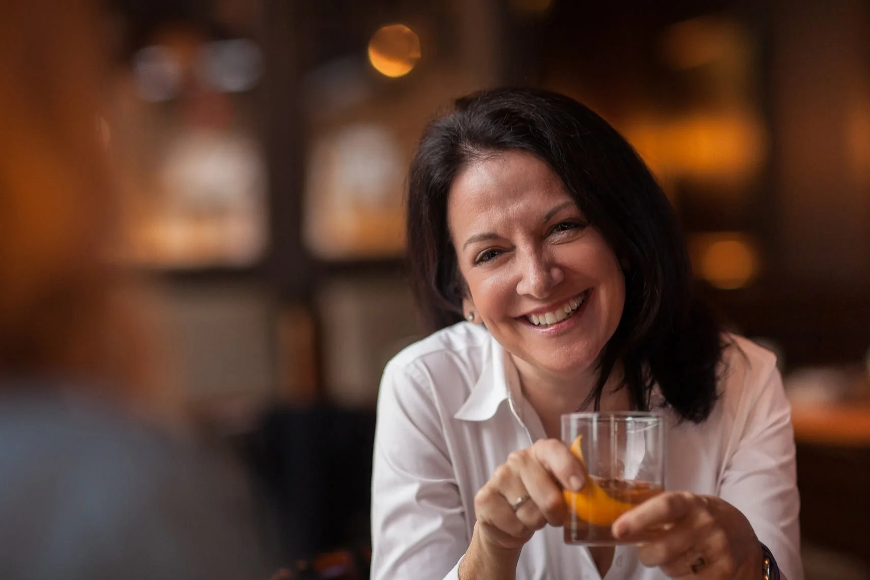 A smiling woman with dark hair and a white shirt holding a glass of whiskey or bourbon in a warmly lit setting, possibly a bar or restaurant.
