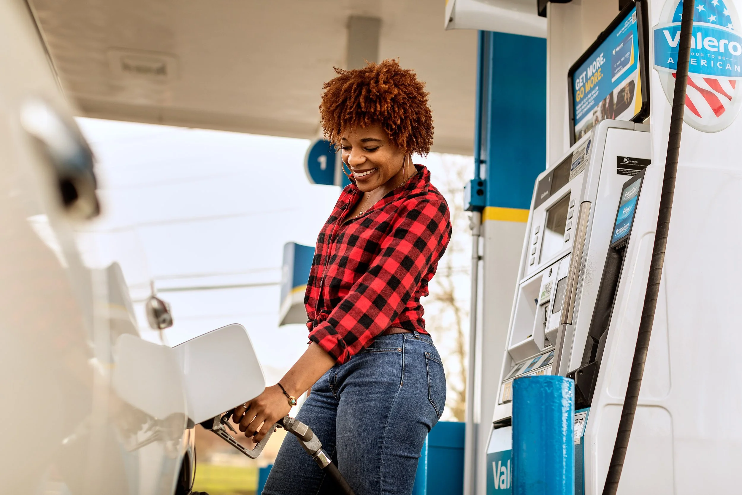 A woman with curly reddish-brown hair in a red and black plaid shirt filling up her car with gas at a Valero gas station.