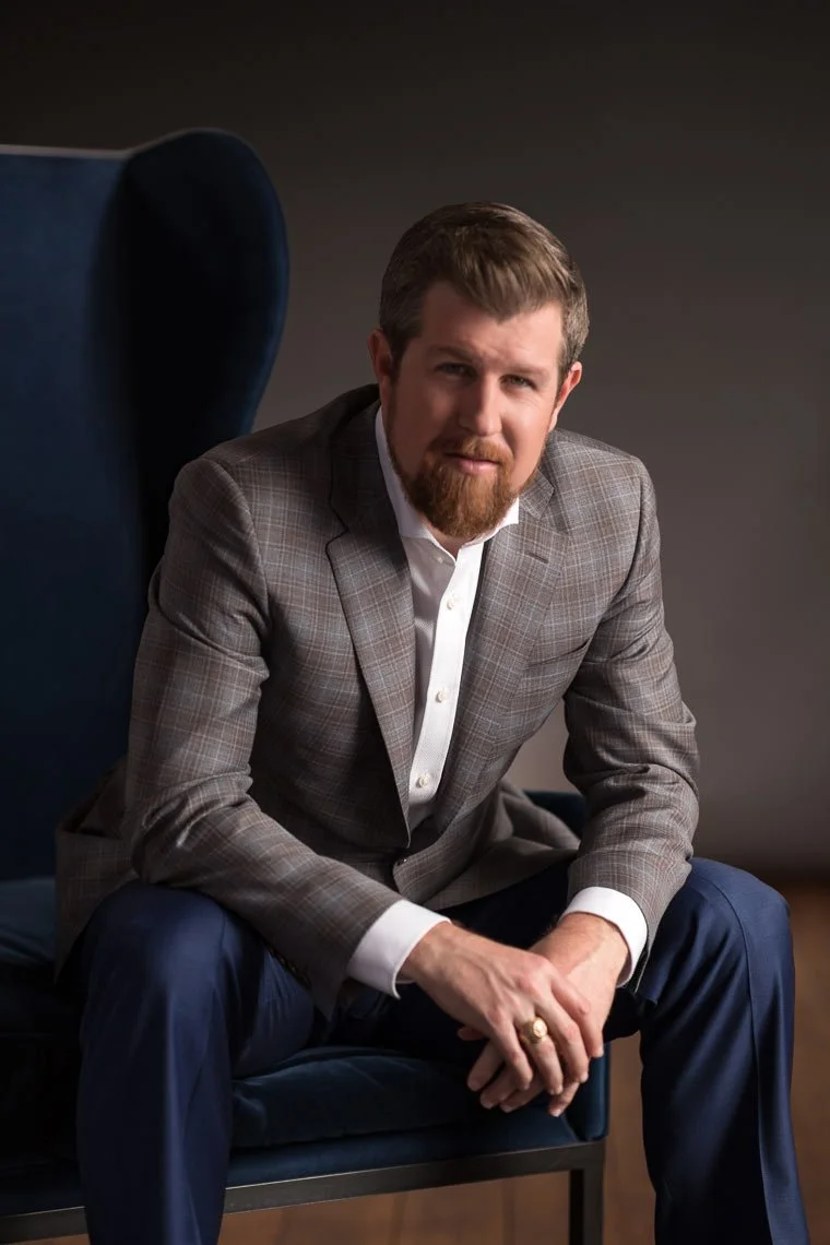 A man with a beard and mustache, wearing a gray checked blazer and navy pants, sitting on a dark blue armchair against a dark background.
