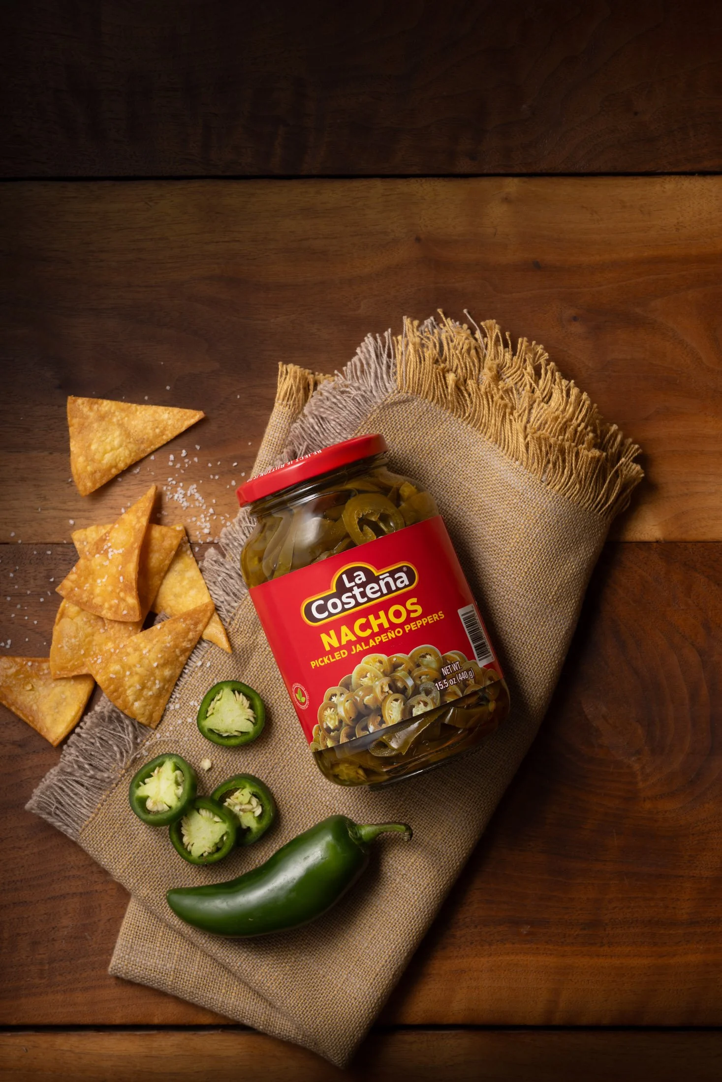 A jar of La Costena pickled jalapeño peppers, sliced jalapeños, scattered salt, tortilla chips, a green jalapeño pepper, and a small section of a green chili pepper on a beige cloth placed on a wooden surface.
