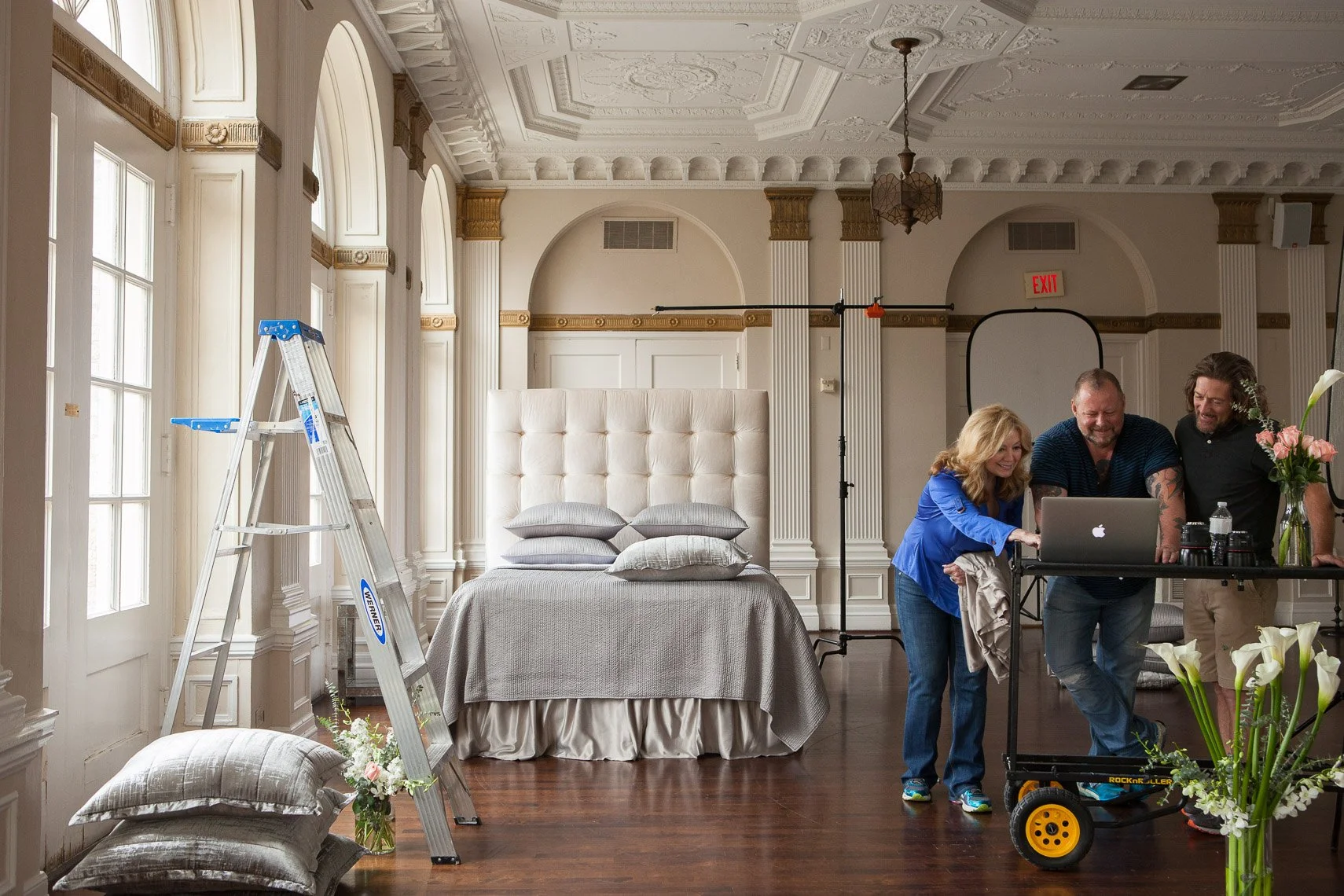 A behind-the-scenes look at a photoshoot in a luxurious room with ornate ceiling and large windows, showing three smiling people gathered around a laptop, with filming equipment and props around them.