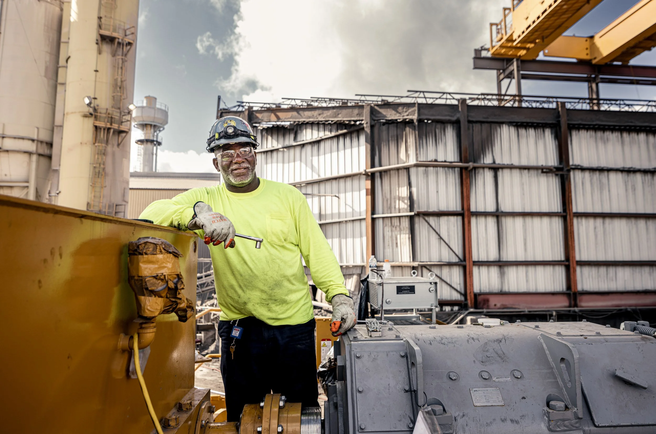 A worker in safety gear stands next to industrial equipment outdoors at a construction or industrial site, with large metal structures and a partly cloudy sky in the background.