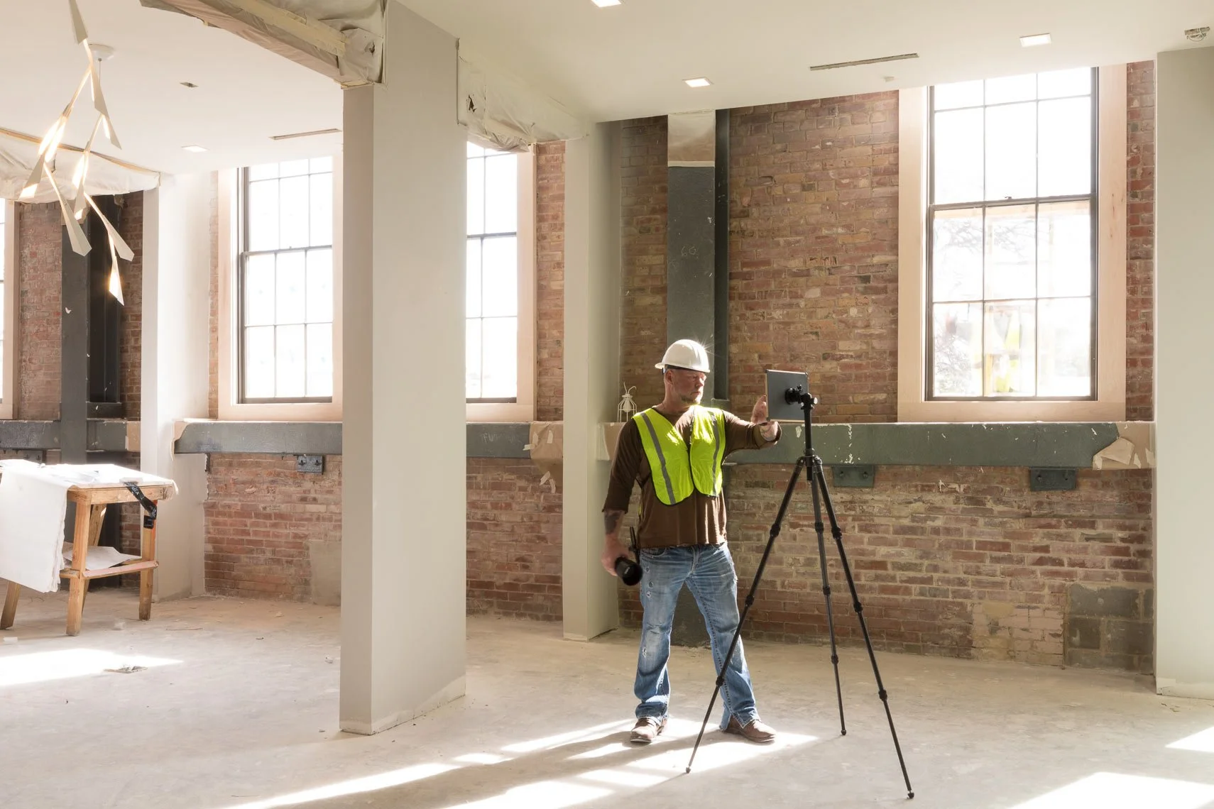 A construction worker wearing a white helmet and neon yellow safety vest taking a photo or video with a tripod-mounted device inside an unfinished building with brick walls and large windows.