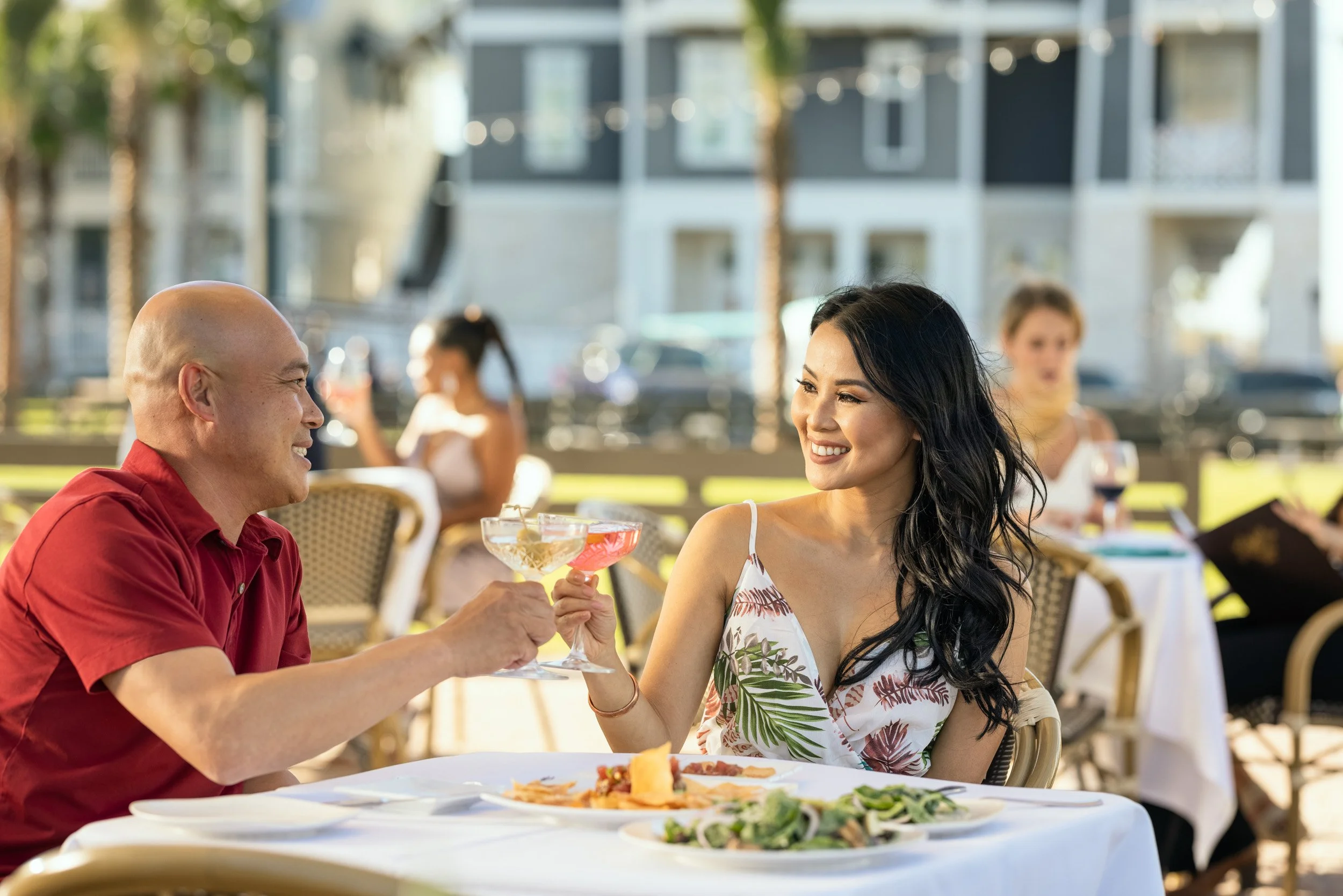 A man and woman smiling and holding cocktails at an outdoor restaurant during daytime. The woman wears a floral dress and the man wears a red shirt. There are other people dining in the background.