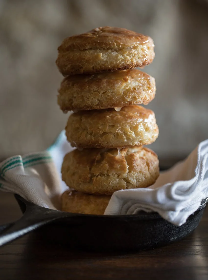 Stack of four freshly baked biscuits in a cast iron skillet with a cloth lining, on a wooden surface.