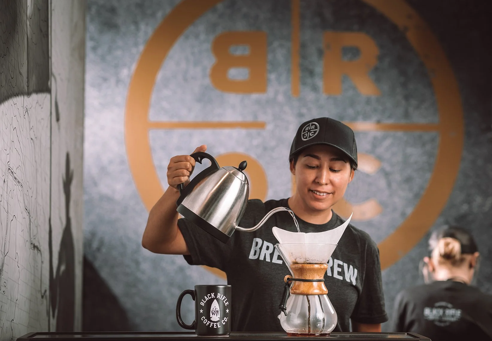 A woman pouring hot water from a kettle into a coffee dripper with a paper filter, on a counter in a coffee shop. She is smiling and wearing a hat and a black t-shirt that says 'Brewing'.